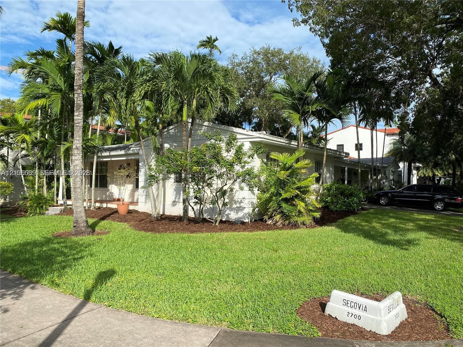 Single-story residence featuring a smooth stucco exterior, covered front porch, manicured lawn, and mature tropical landscaping