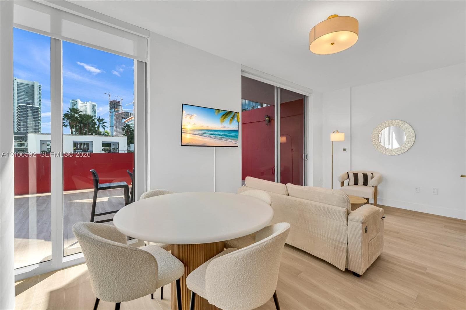 Floor-to-ceiling sliding glass doors, light wood-finish flooring, recessed drum lighting, and expansive private balcony with red-toned exterior walls