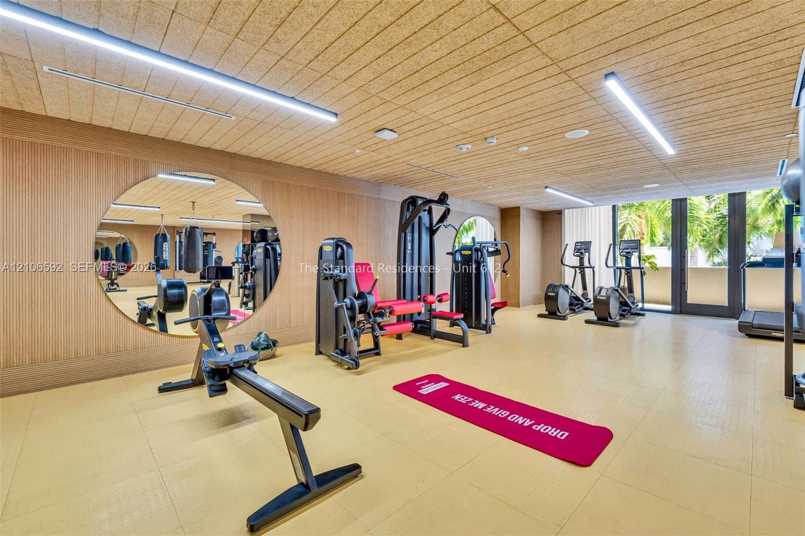Fitness center featuring wood-paneled walls, a large circular wall-mounted mirror, linear LED lighting, and glass doors with exterior foliage