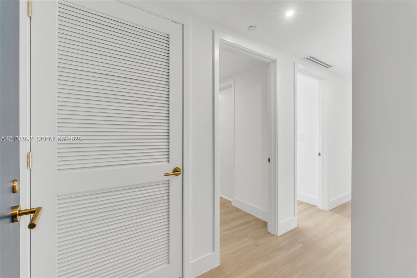 Interior hallway featuring wood-finish flooring, bright white walls, and white louvered doors with satin brass hardware