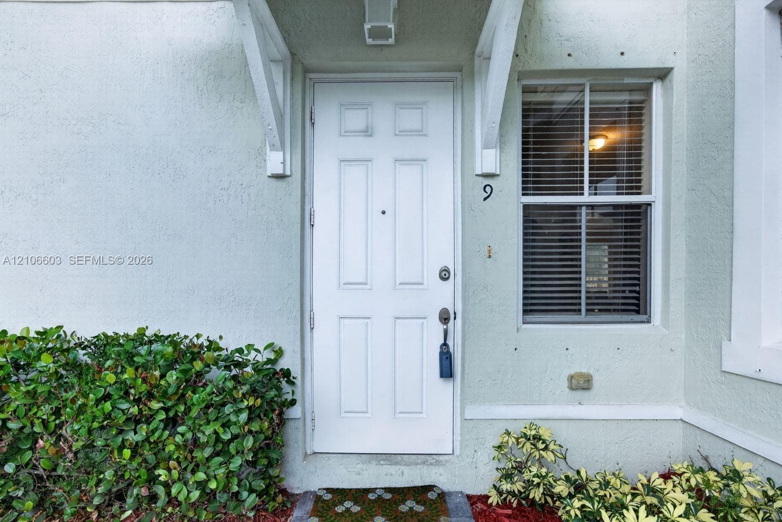 Entryway featuring a six-panel white door, arched wall-mounted portico, textured stucco exterior, and multi-paned window with horizontal blinds