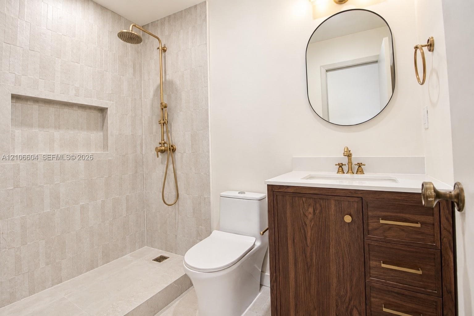 Bathroom featuring a wood-finish vanity with a stone countertop, wall-mounted oval mirror, brass-toned fixtures, walk-in shower with vertical tile surround, and a built-in shower niche
