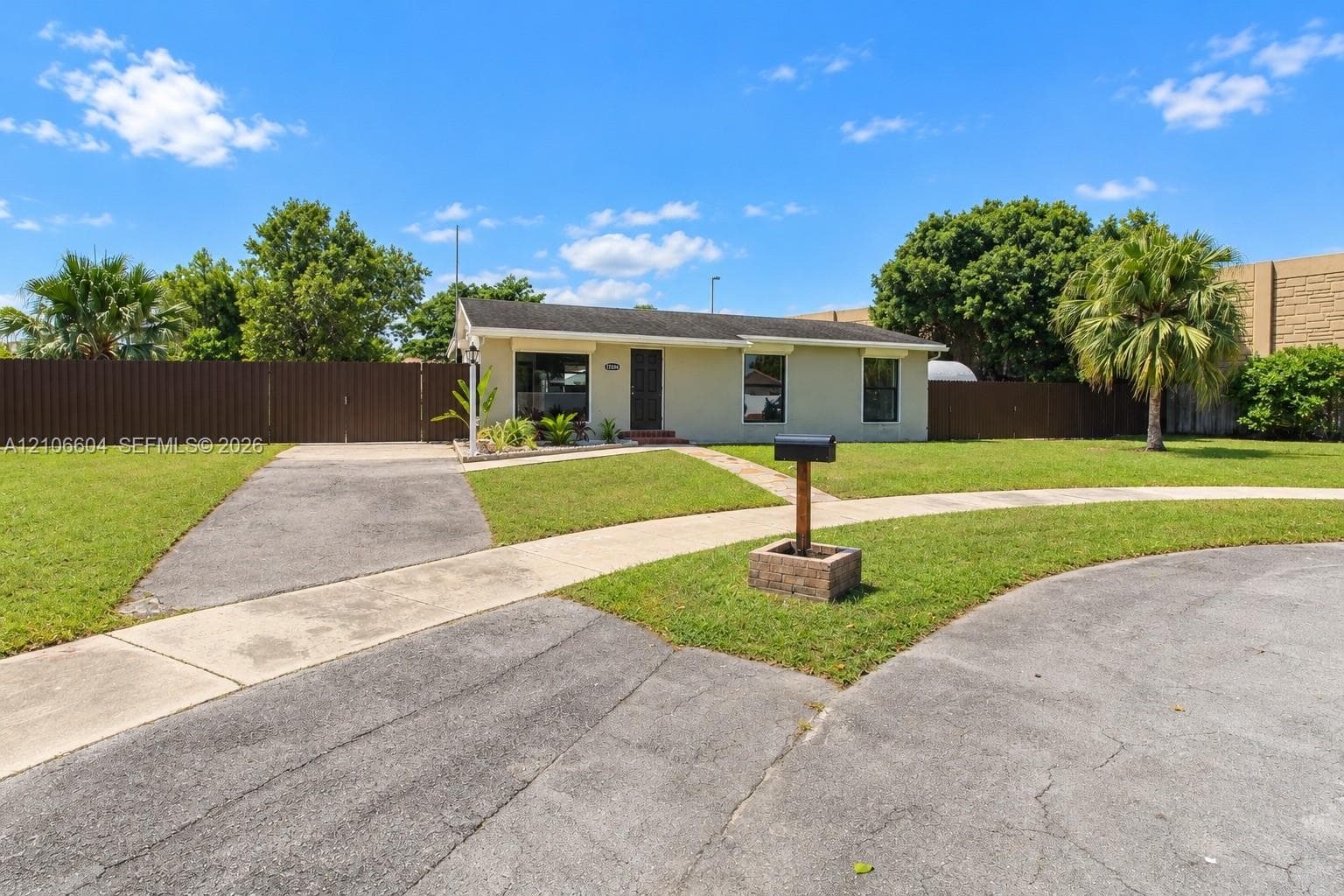 Single-story exterior featuring a light-toned stucco facade, asphalt shingle roof, expansive front lawn, paved driveway, and privacy fencing