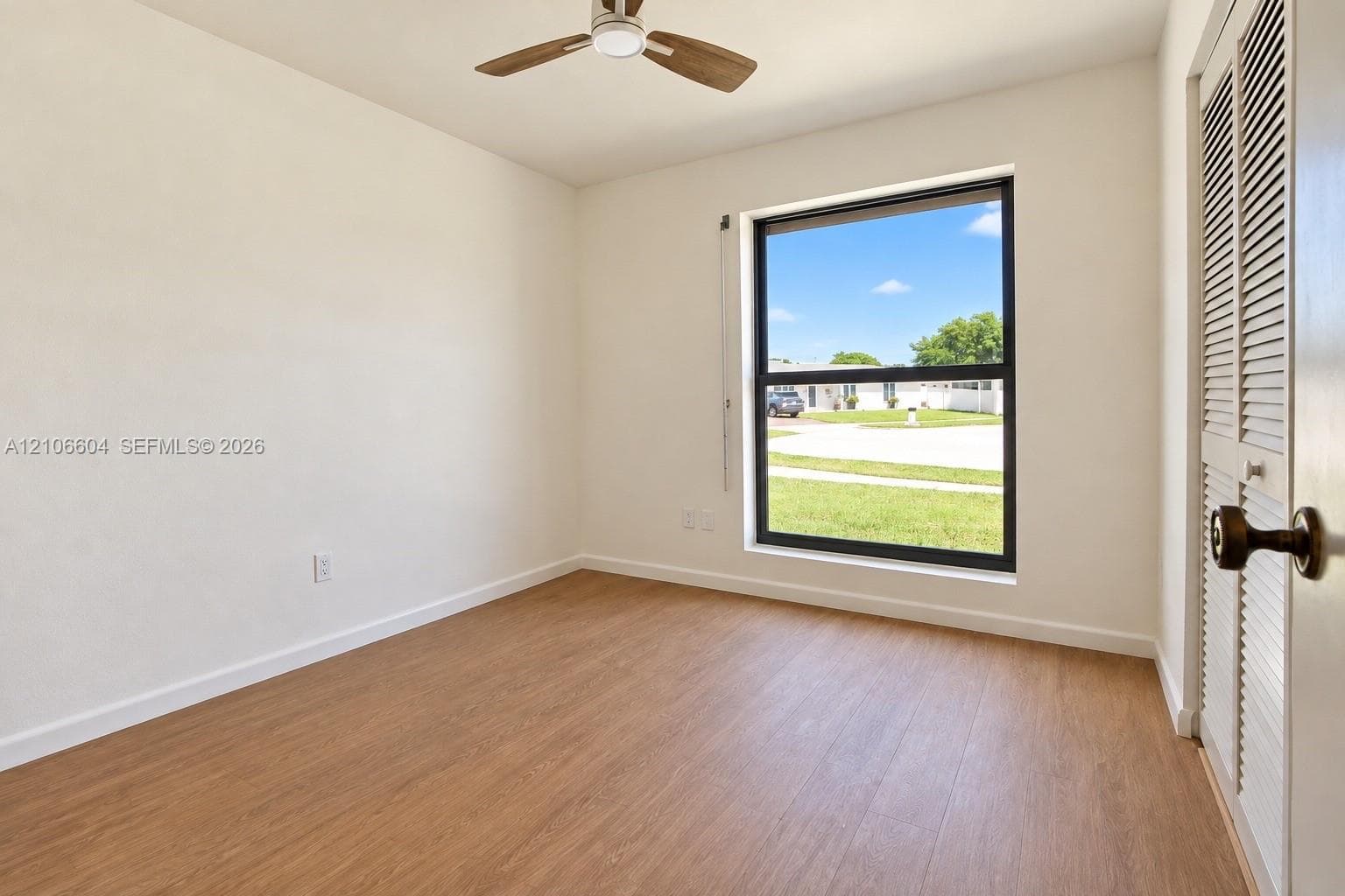 Room featuring wood-finish flooring, a ceiling fan, a large window with a dark frame, and louvered bi-fold closet doors