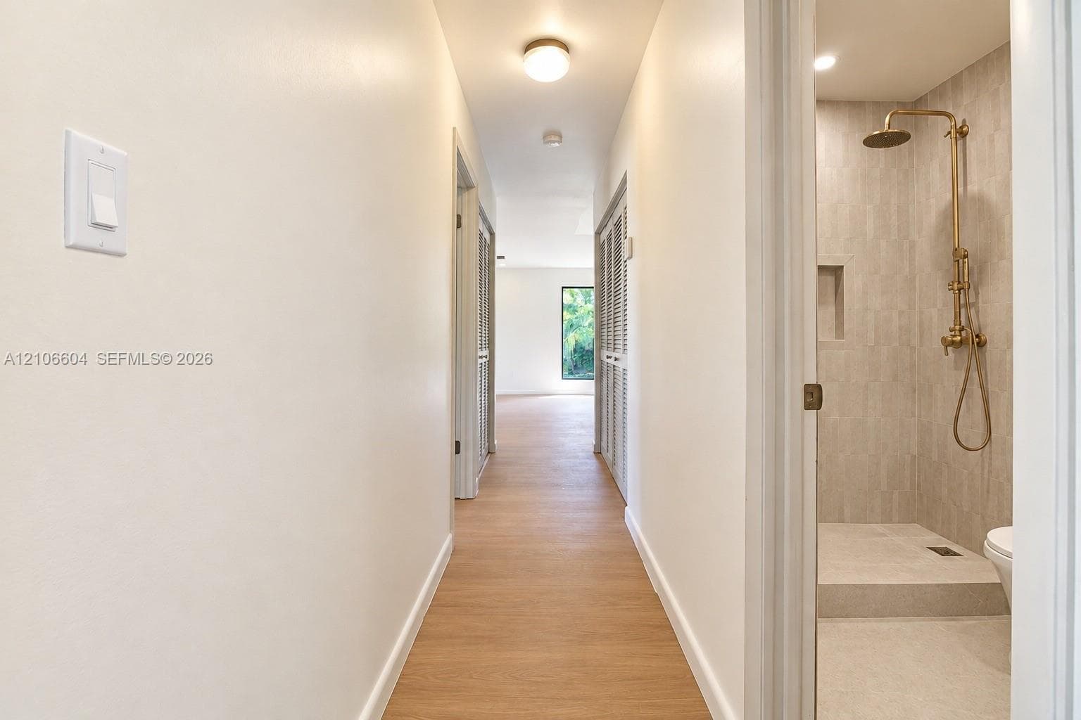 Hallway featuring wood-finish flooring and flush-mount lighting, with a bathroom entry showcasing a tiled walk-in shower, built-in wall niche, and brass-finish fixtures
