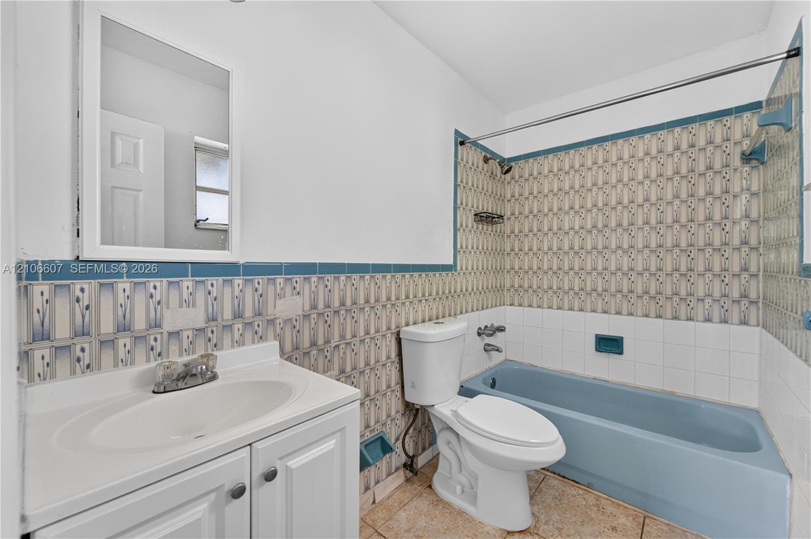 Bathroom featuring a vanity with integrated sink, rectangular wall-mounted mirror, blue tub with tiled surround, and ceramic tile flooring