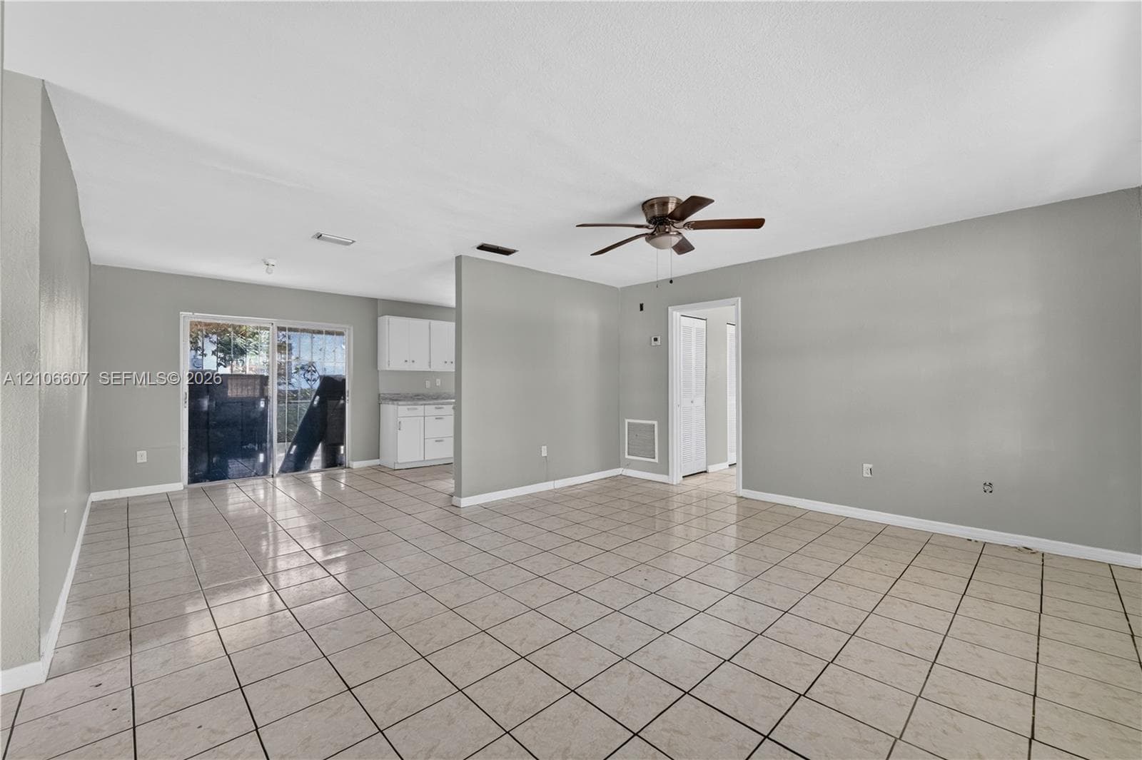 Open-concept interior featuring light-toned tile flooring, ceiling fan with light fixture, sliding glass doors, and white cabinetry