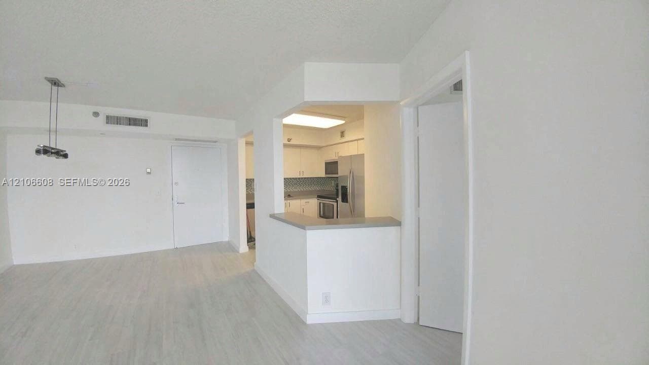 Open-concept interior featuring light-toned wood-finish flooring, a kitchen pass-through counter, neutral cabinetry, patterned tile backsplash, and a modern multi-light pendant fixture