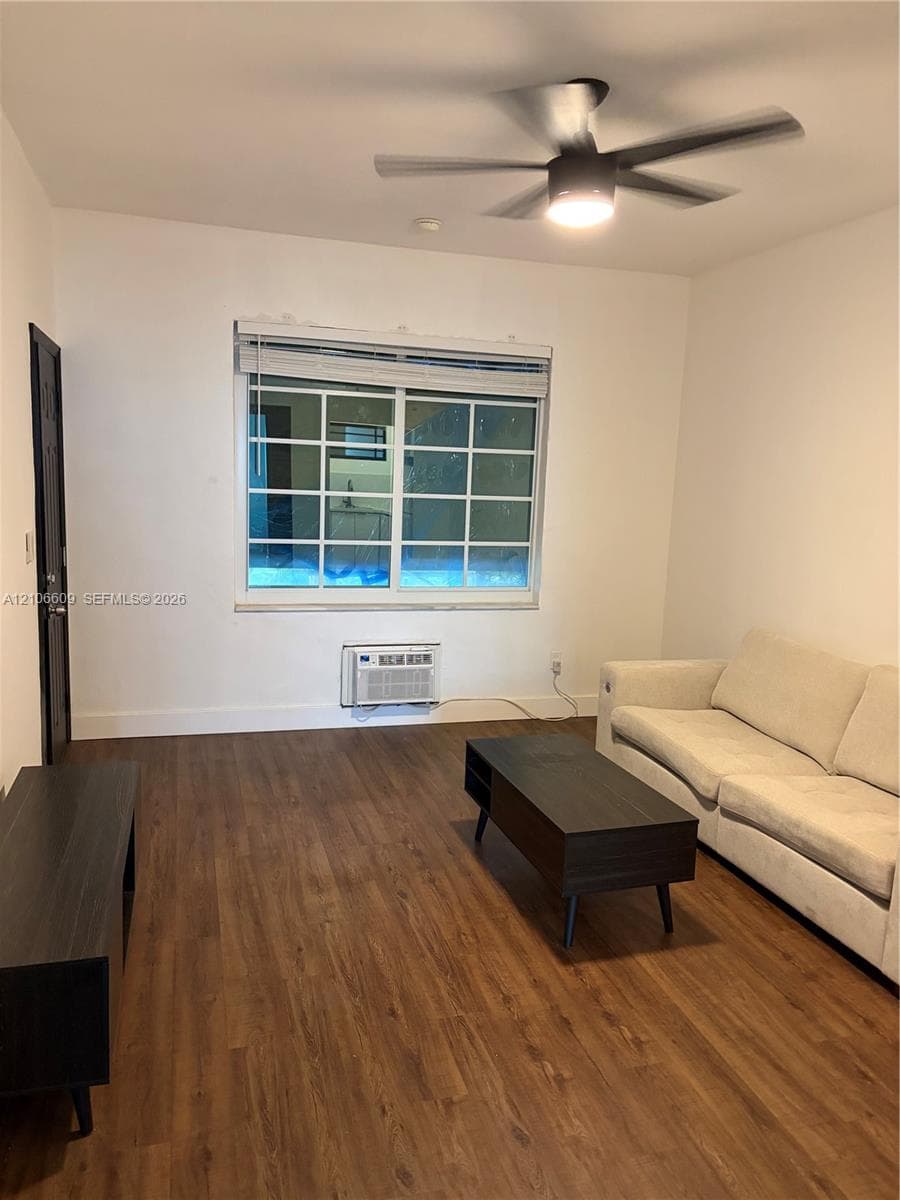 Wood-finish flooring, multi-pane window with horizontal blinds, integrated wall-mounted air conditioning unit, and a central ceiling fan with light fixture