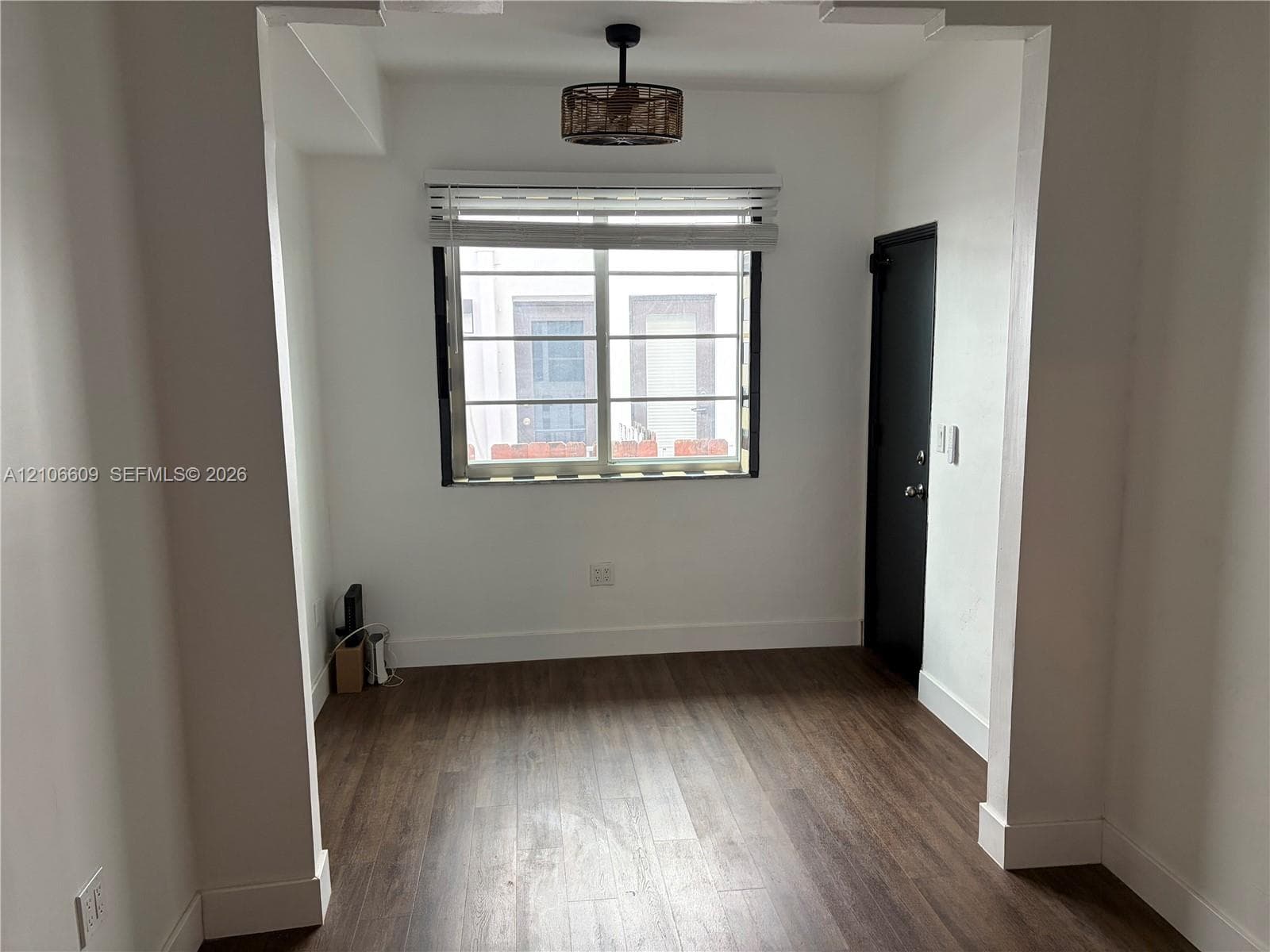 Room featuring wood-finish flooring, a drum-style ceiling light fixture, a single-hung window with horizontal blinds, and a solid-panel door with dark hardware