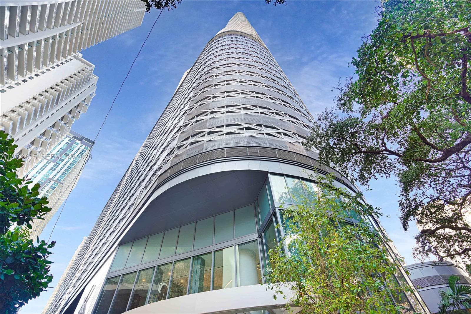 High-rise architectural structure featuring a curved glass facade, geometric metal lattice cladding, and floor-to-ceiling glass paneling at the street level