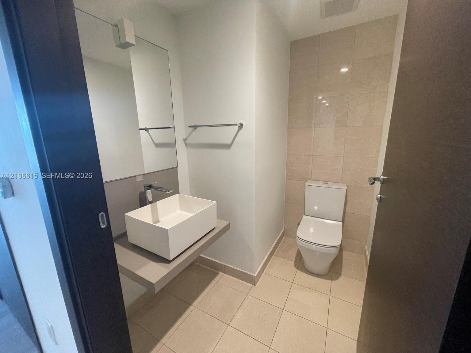 Floating vanity with rectangular vessel sink, wall-mounted faucet, frameless mirror, and neutral floor tile featuring a tiled accent wall