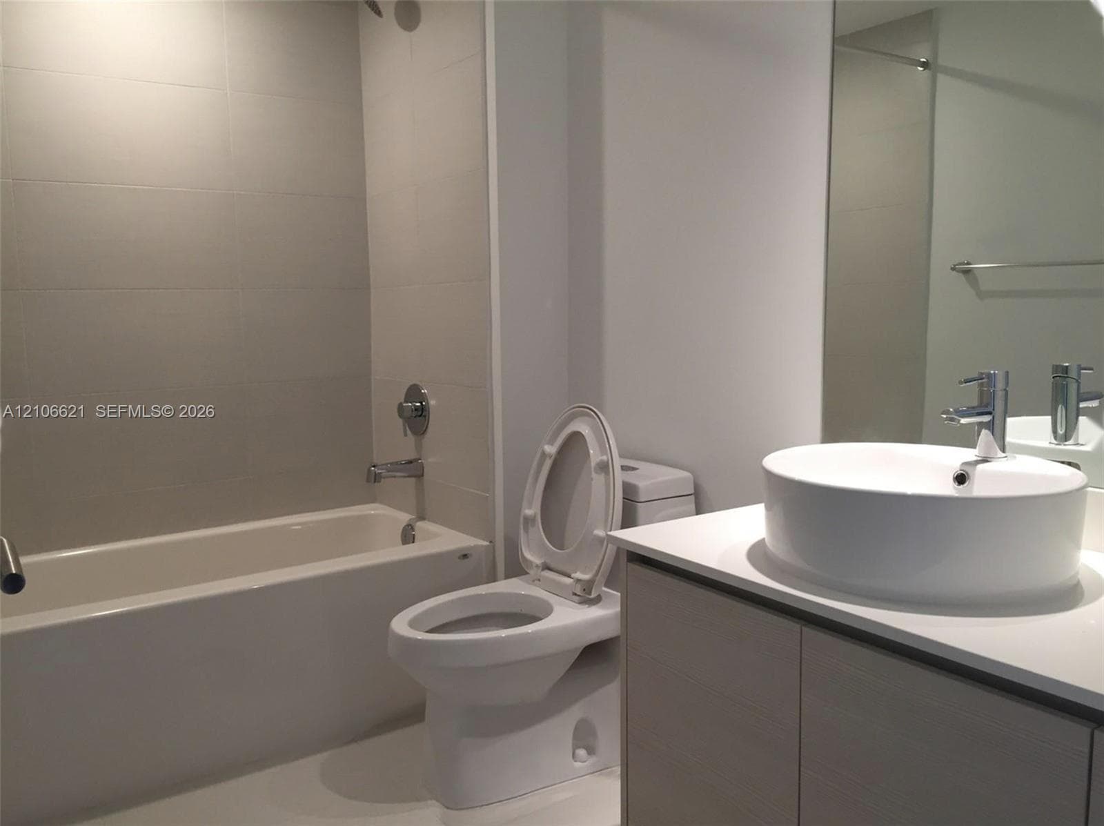 Bathroom featuring a vanity with a rectangular vessel sink, white solid surface countertop, wood-finish cabinetry, a bathtub with full-height tile surround, and a large-format mirror