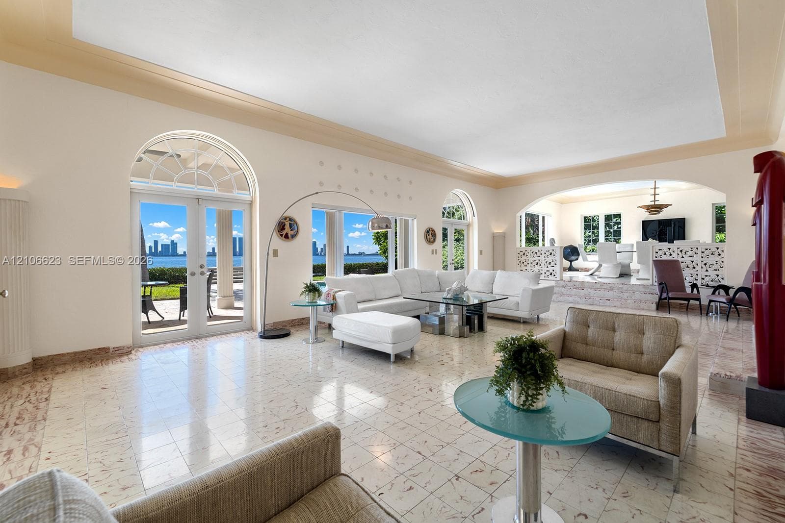 Open-concept interior featuring light-toned stone flooring, arched glass double doors, elevated platform area with decorative latticework, and recessed tray ceiling detailing