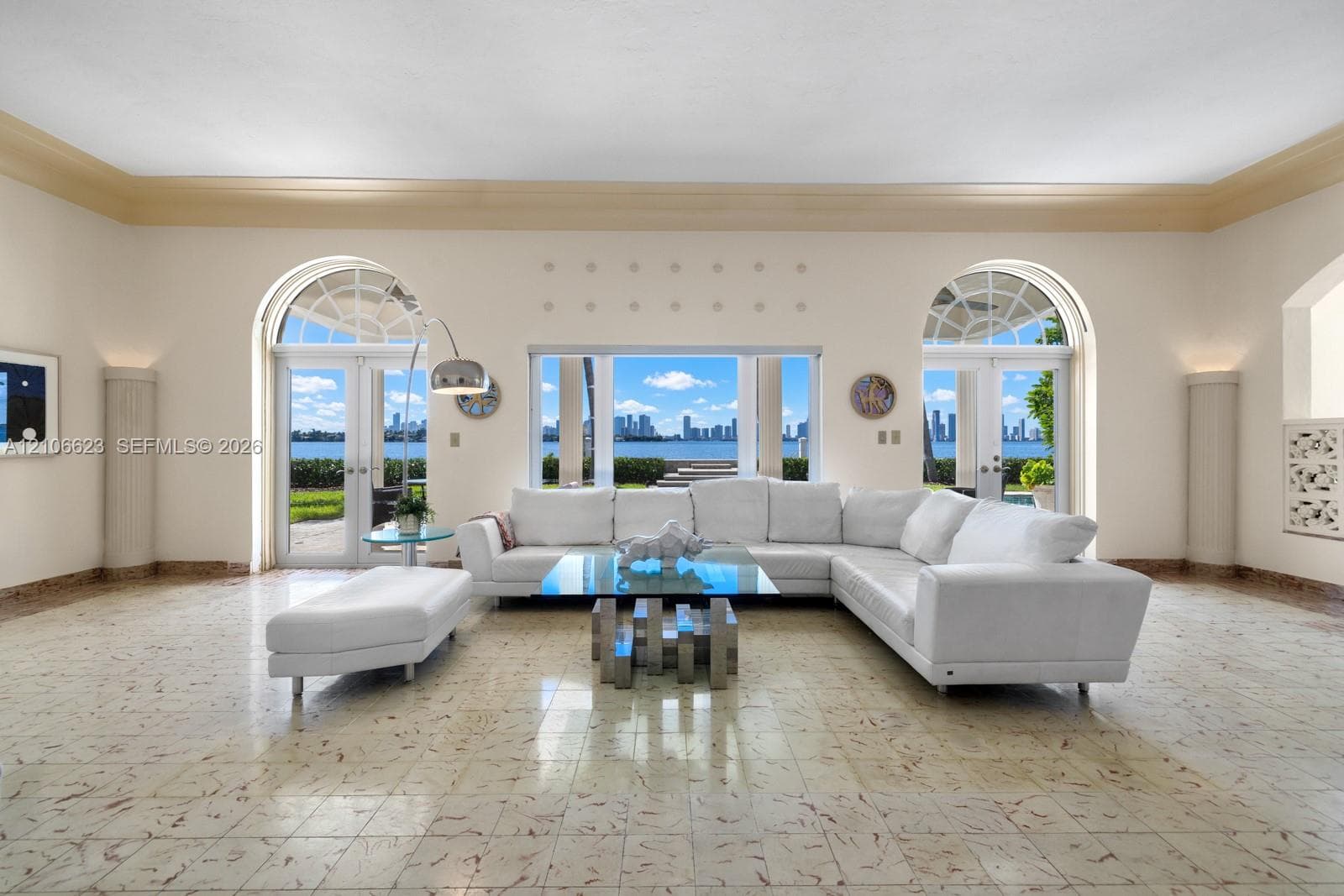 Living area featuring stone flooring, arched glass double doors, large central picture window, decorative crown molding, and recessed wall columns