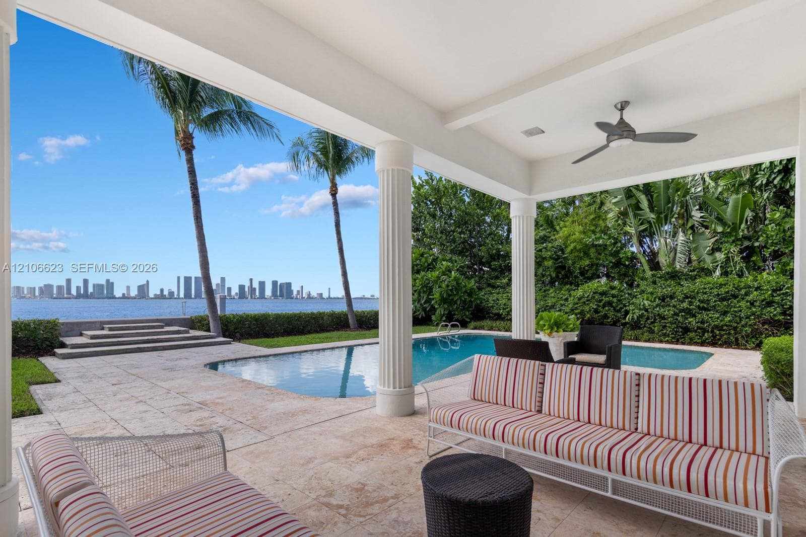 Covered patio featuring stone tile flooring, structural columns, and a ceiling-mounted fan, overlooking an in-ground swimming pool and waterfront with skyline backdrop
