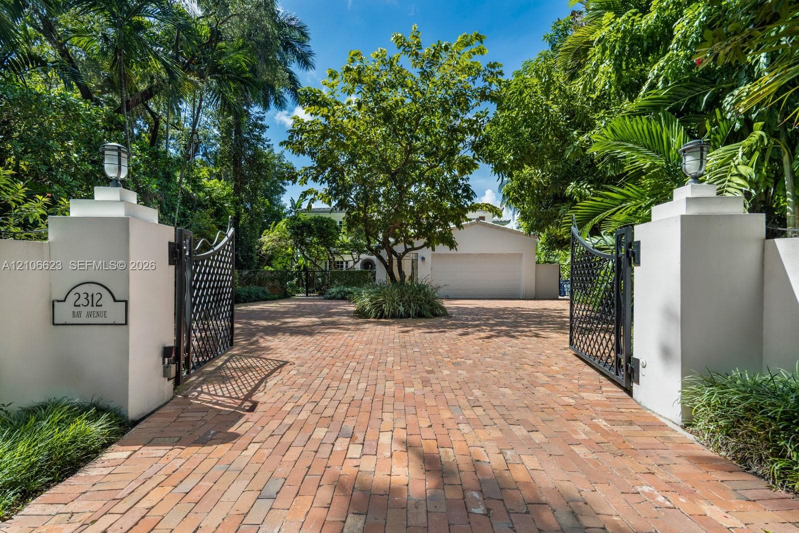 Gated entrance featuring stucco pillars with crown lighting, decorative metal gates, brick paver driveway, and mature landscaping