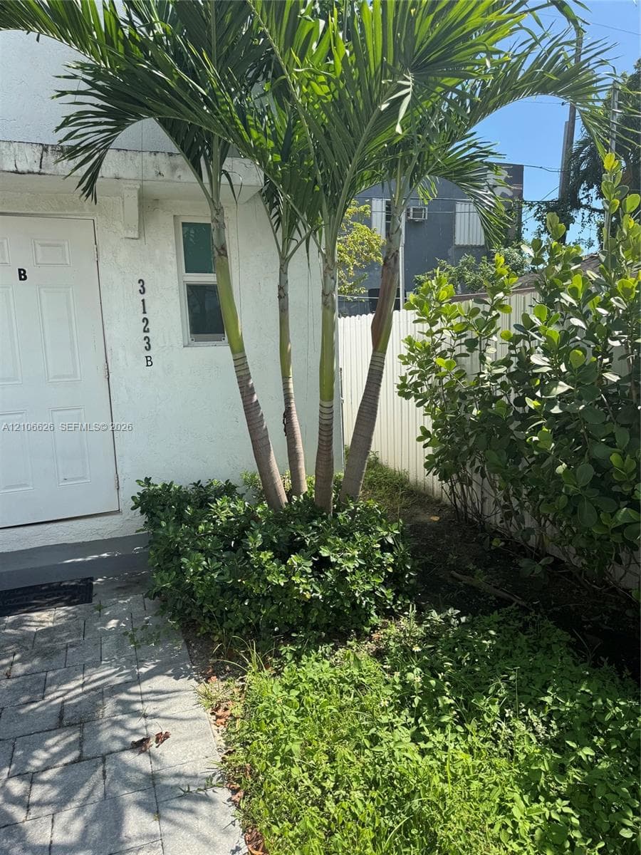 Stucco exterior featuring a paved walkway, mature palm trees, and perimeter fencing
