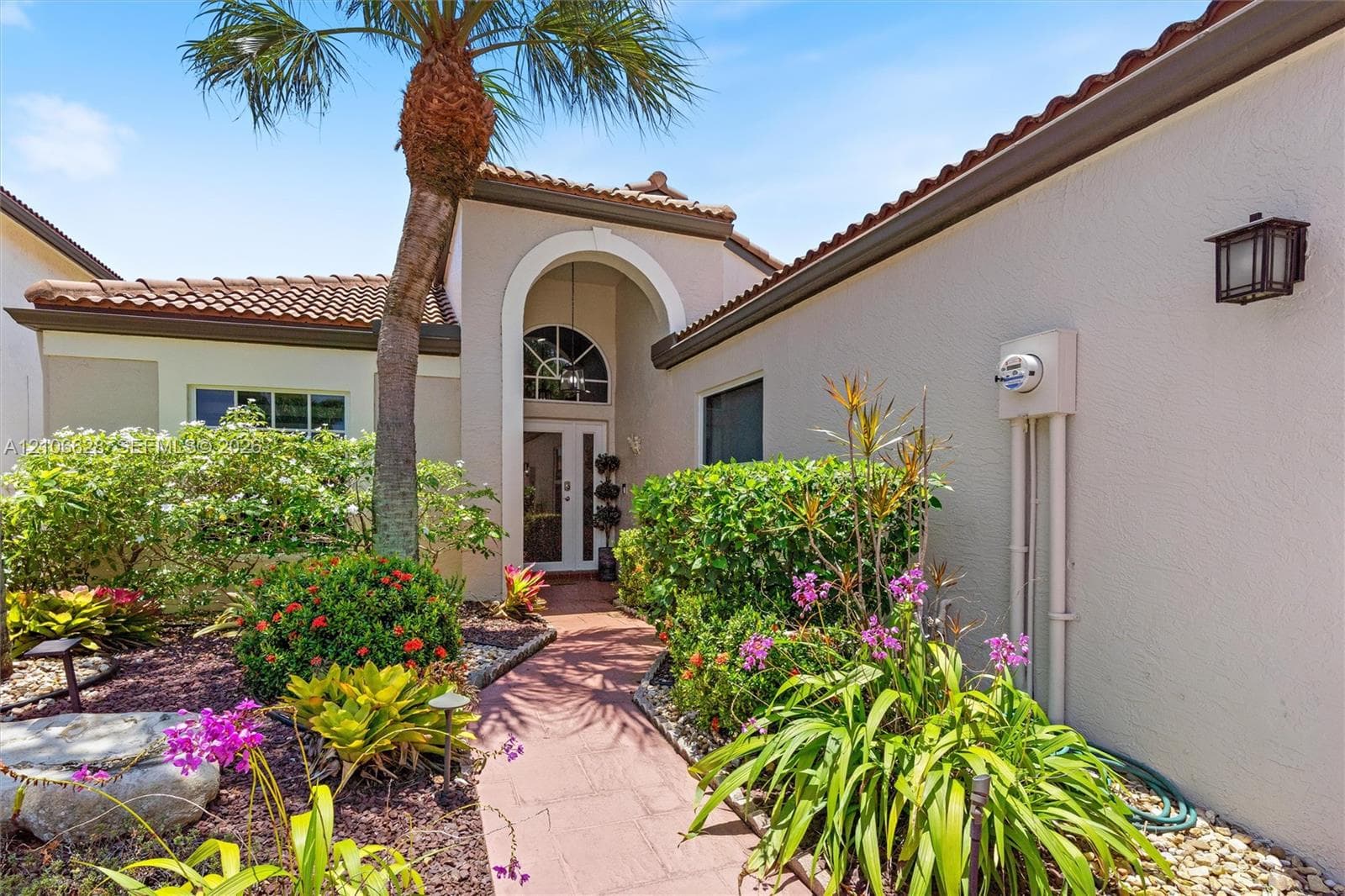 Stucco exterior featuring a arched entryway, tile roof, and a paved walkway surrounded by mature landscaping