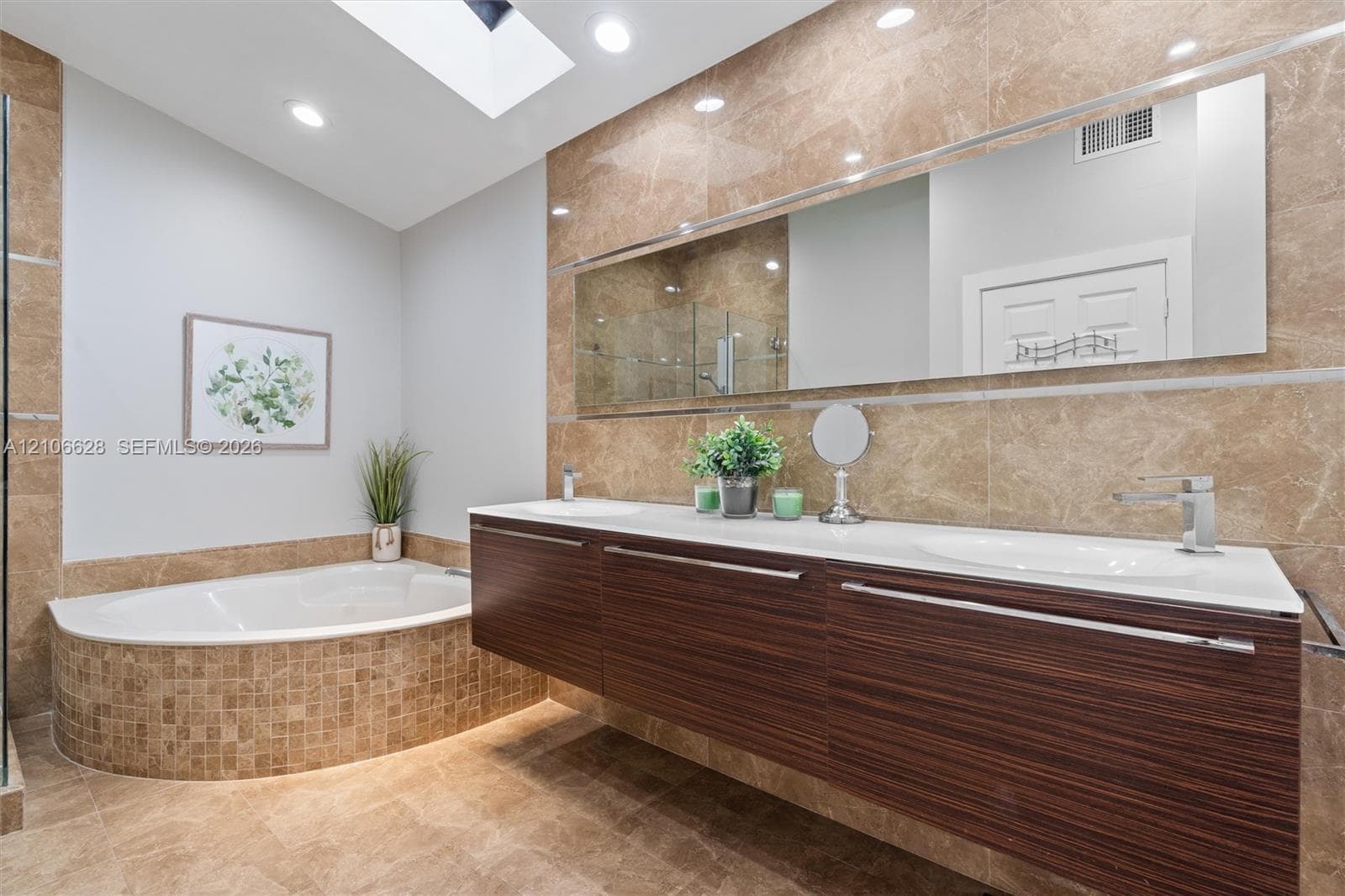 Floating vanity with dual-integrated sinks, wood-finish cabinetry, full-width wall mirror, oversized soaking tub with mosaic tile surround, and skylight
