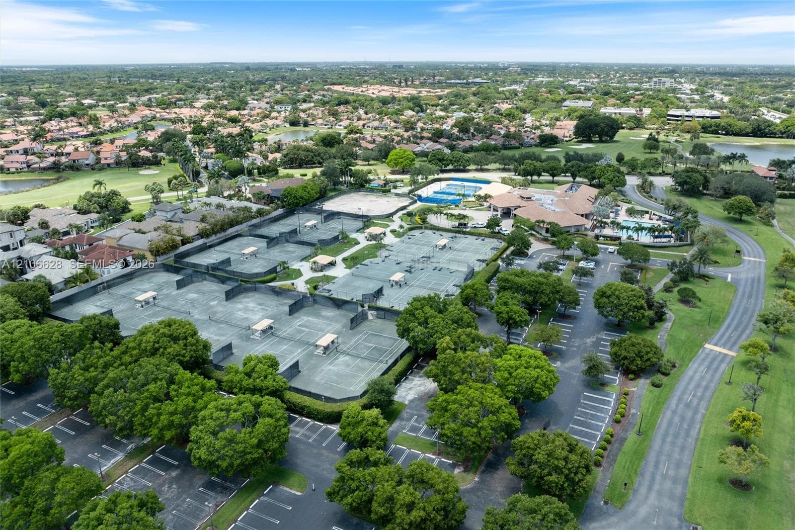 Aerial perspective of a recreation complex featuring multiple tennis courts, a swimming pool, a clubhouse building, and paved parking areas