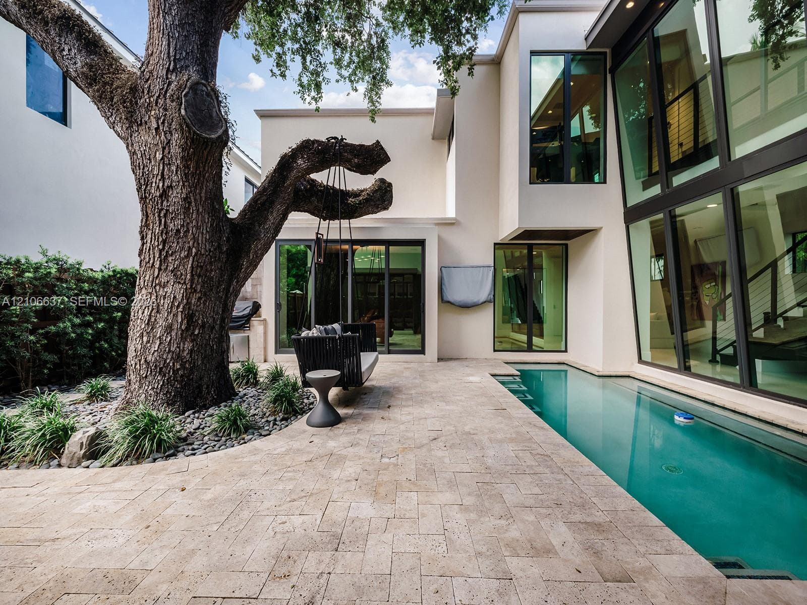 Modern architectural exterior featuring expansive floor-to-ceiling glass windows, a custom geometric lap pool, herringbone-patterned stone decking, and a mature specimen tree