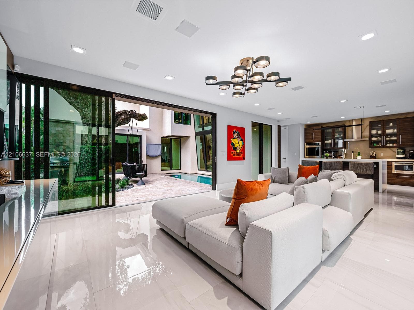 Open-concept interior featuring large-format polished tile flooring, recessed ceiling lighting, a contemporary multi-arm chandelier, and floor-to-ceiling sliding glass doors revealing a paved patio area