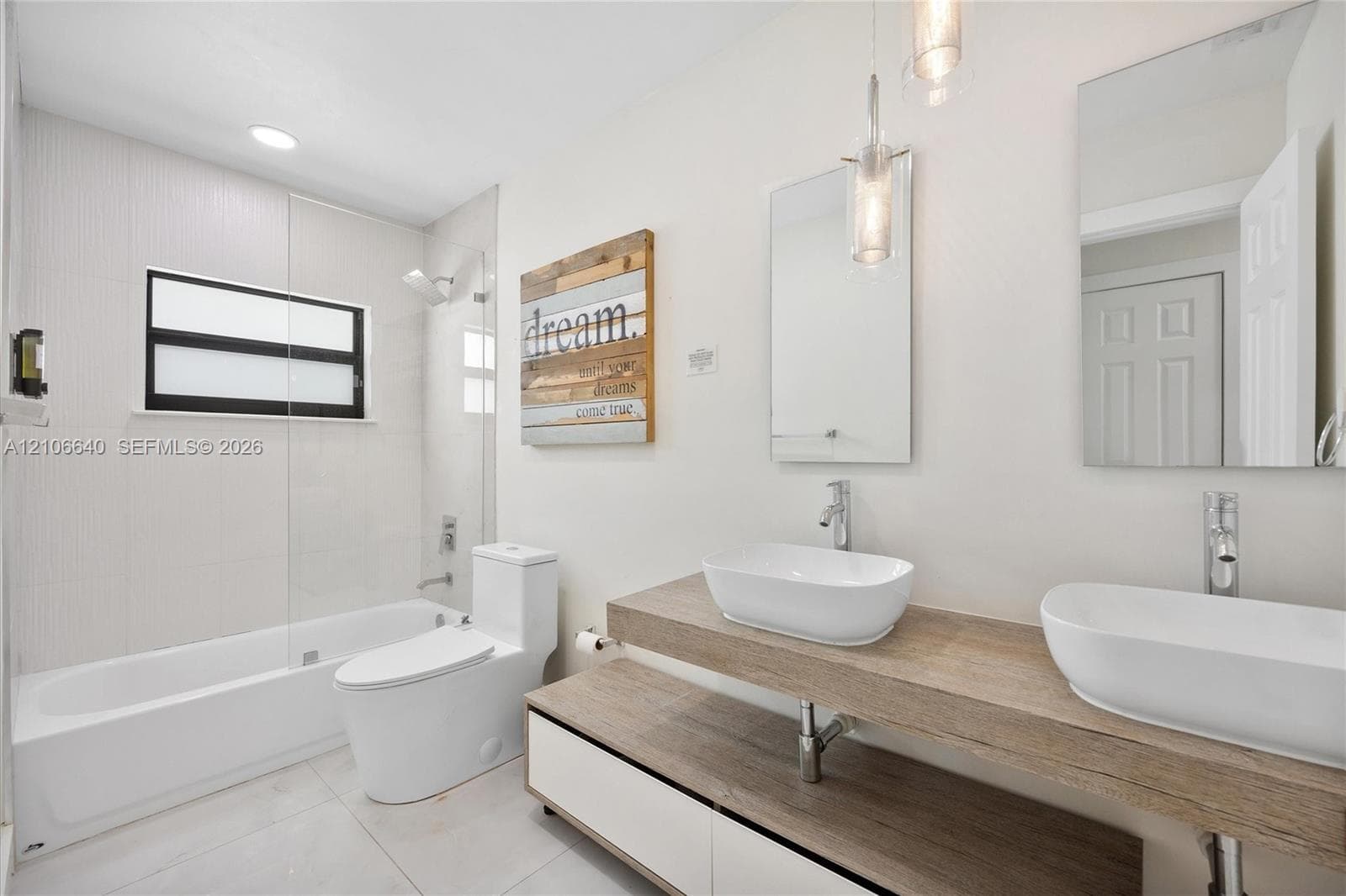 Dual vessel sinks with chrome faucets, wood-finish vanity with integrated storage, full-sized bathtub with glass enclosure, white tile flooring, and recessed lighting