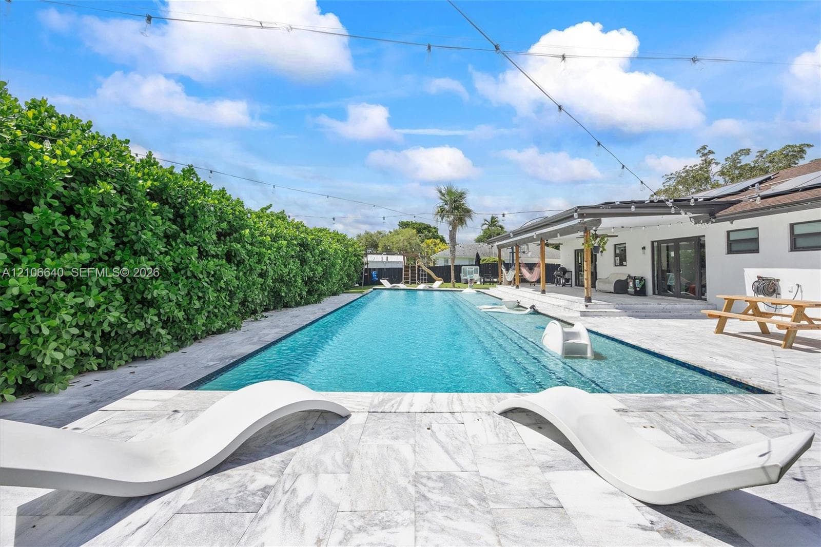 Expansive outdoor living area featuring a rectangular swimming pool, large patio with light-toned pavers, and a covered patio structure