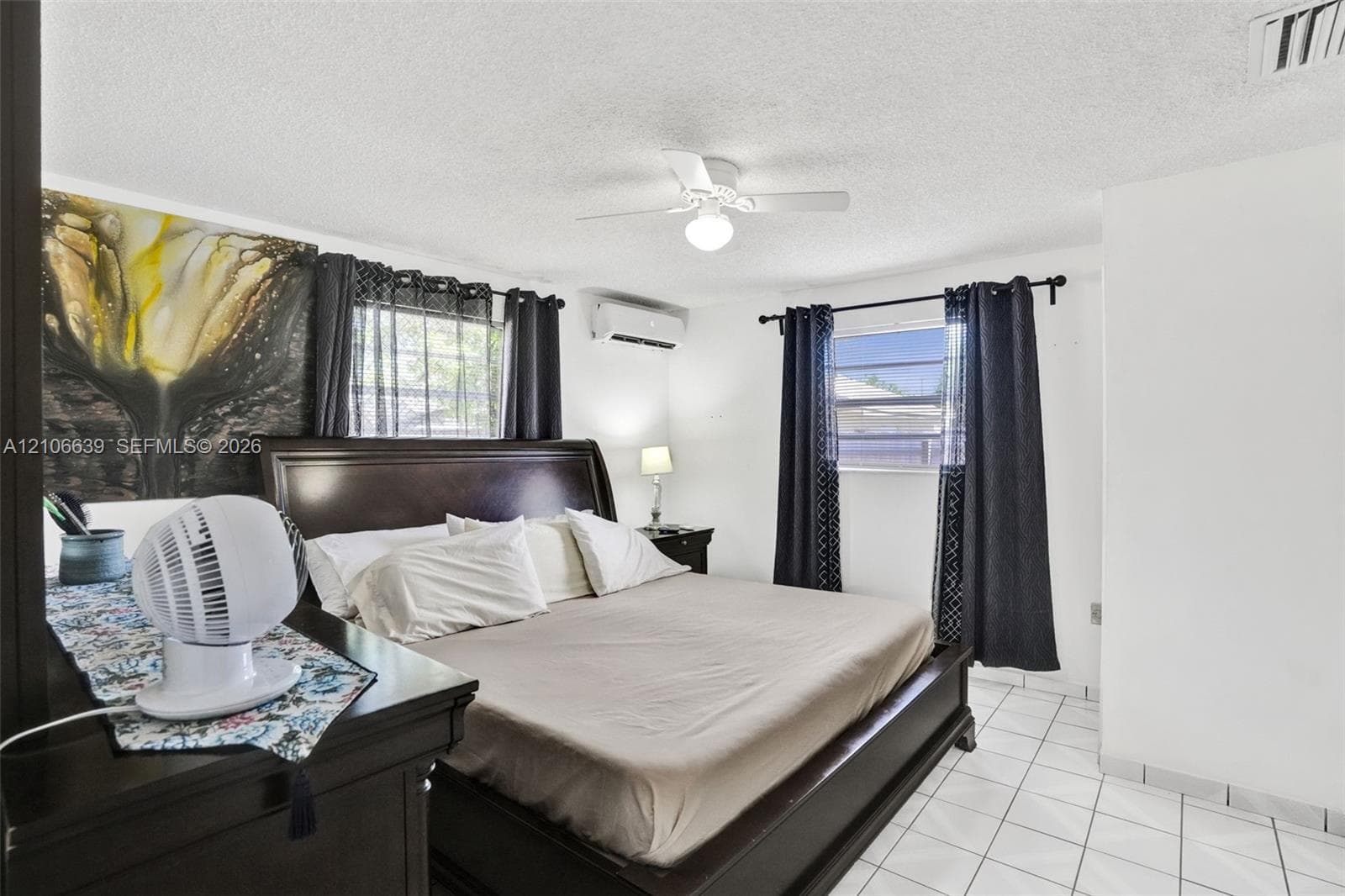 Bedroom featuring white tile flooring, ceiling fan with integrated light fixture, wall-mounted air conditioning unit, and dual windows