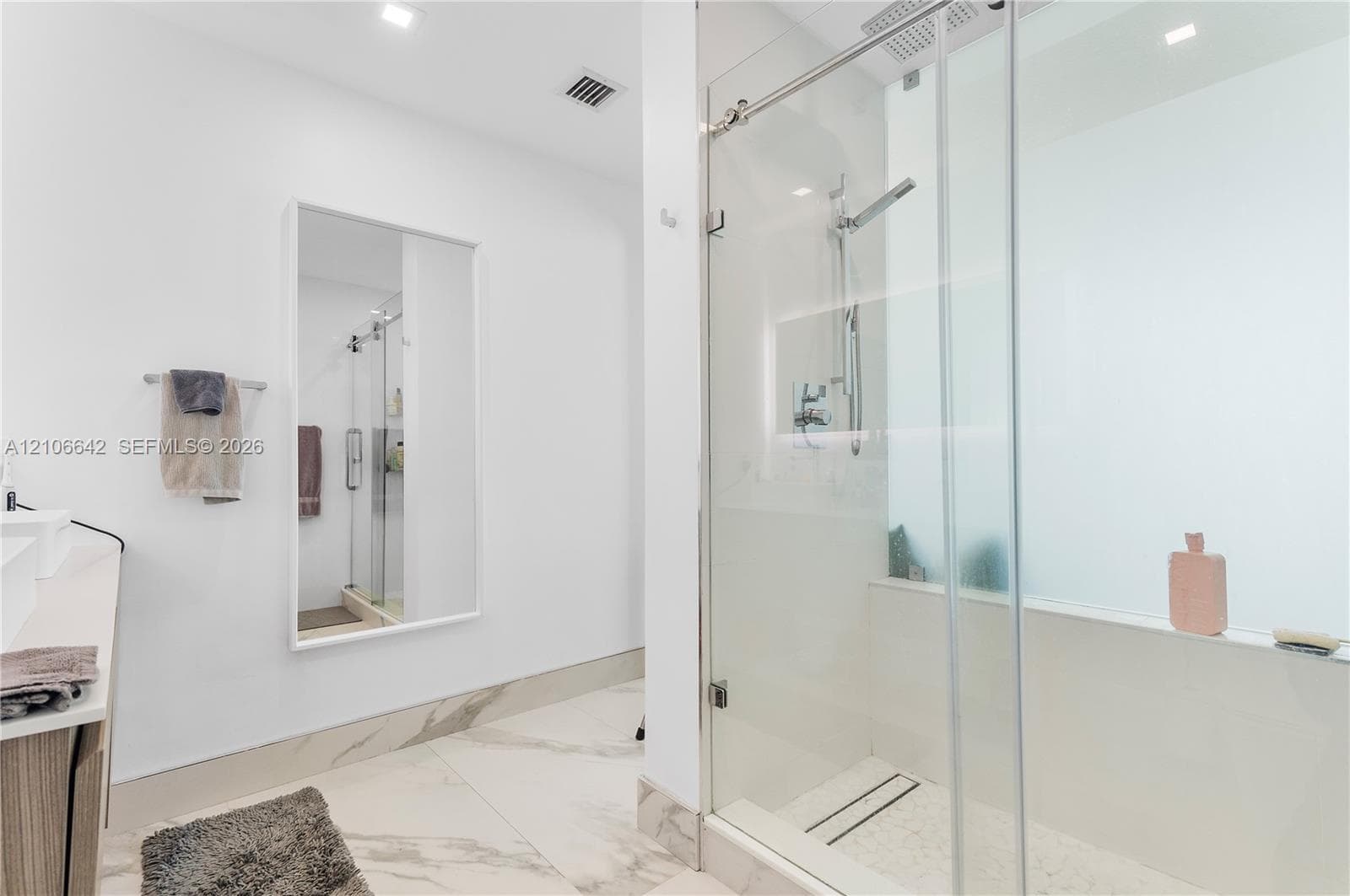 Walk-in shower with a glass enclosure, wall-mounted rainfall showerhead, stone-finish tile flooring, recessed ceiling lighting, and a large wall-mounted mirror