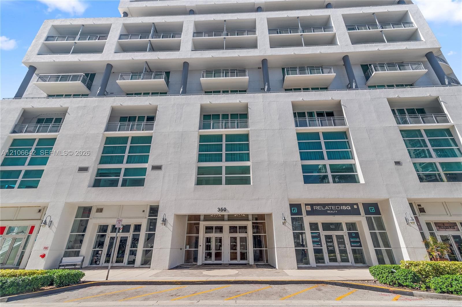 Multi-story concrete exterior featuring floor-to-ceiling glass windows, private balconies with metal railings, and a recessed ground-level entryway