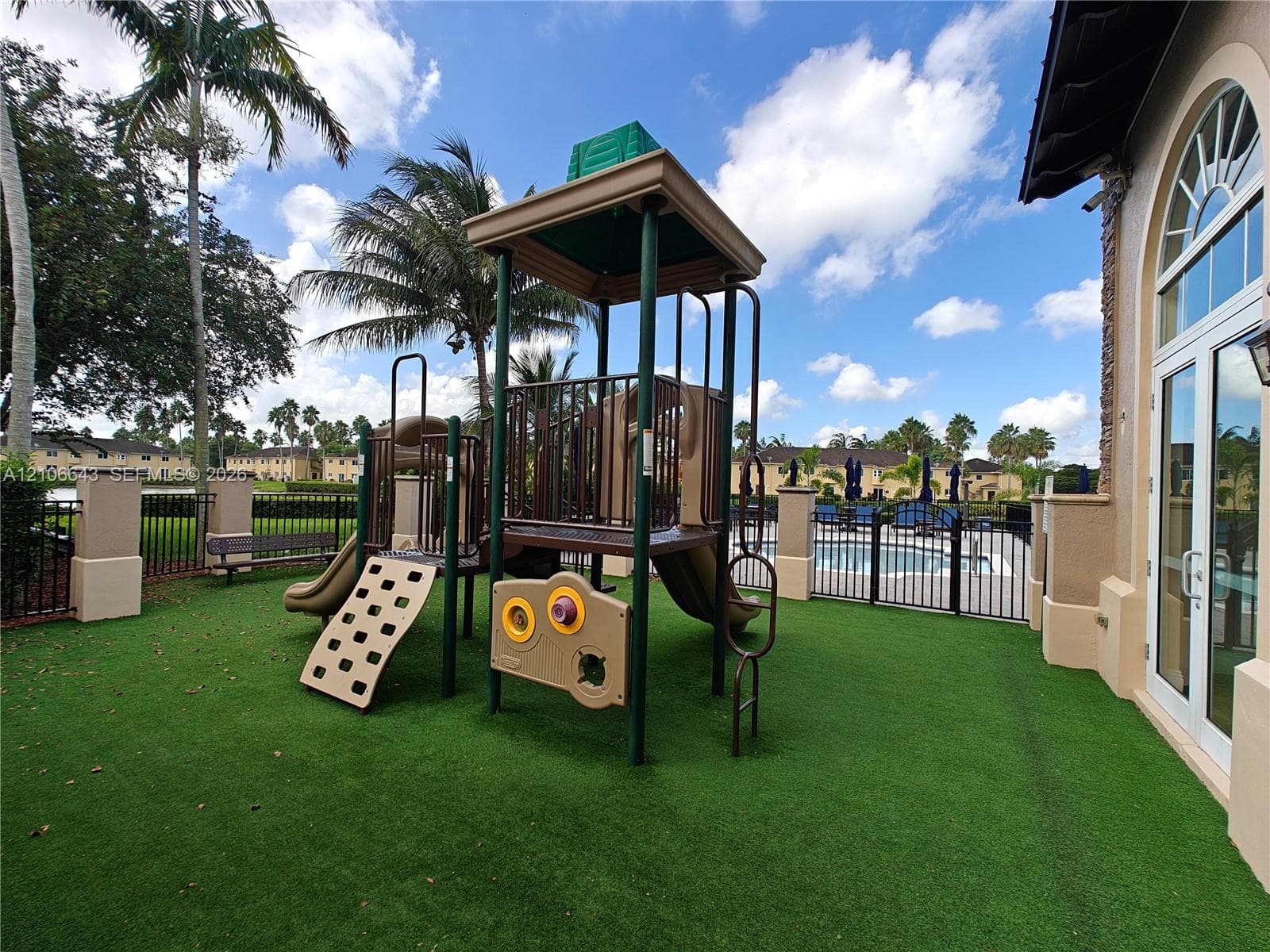 Outdoor recreational area featuring an artificial turf surface, a permanent playground structure, a black metal perimeter fence, and direct exterior access to a swimming pool