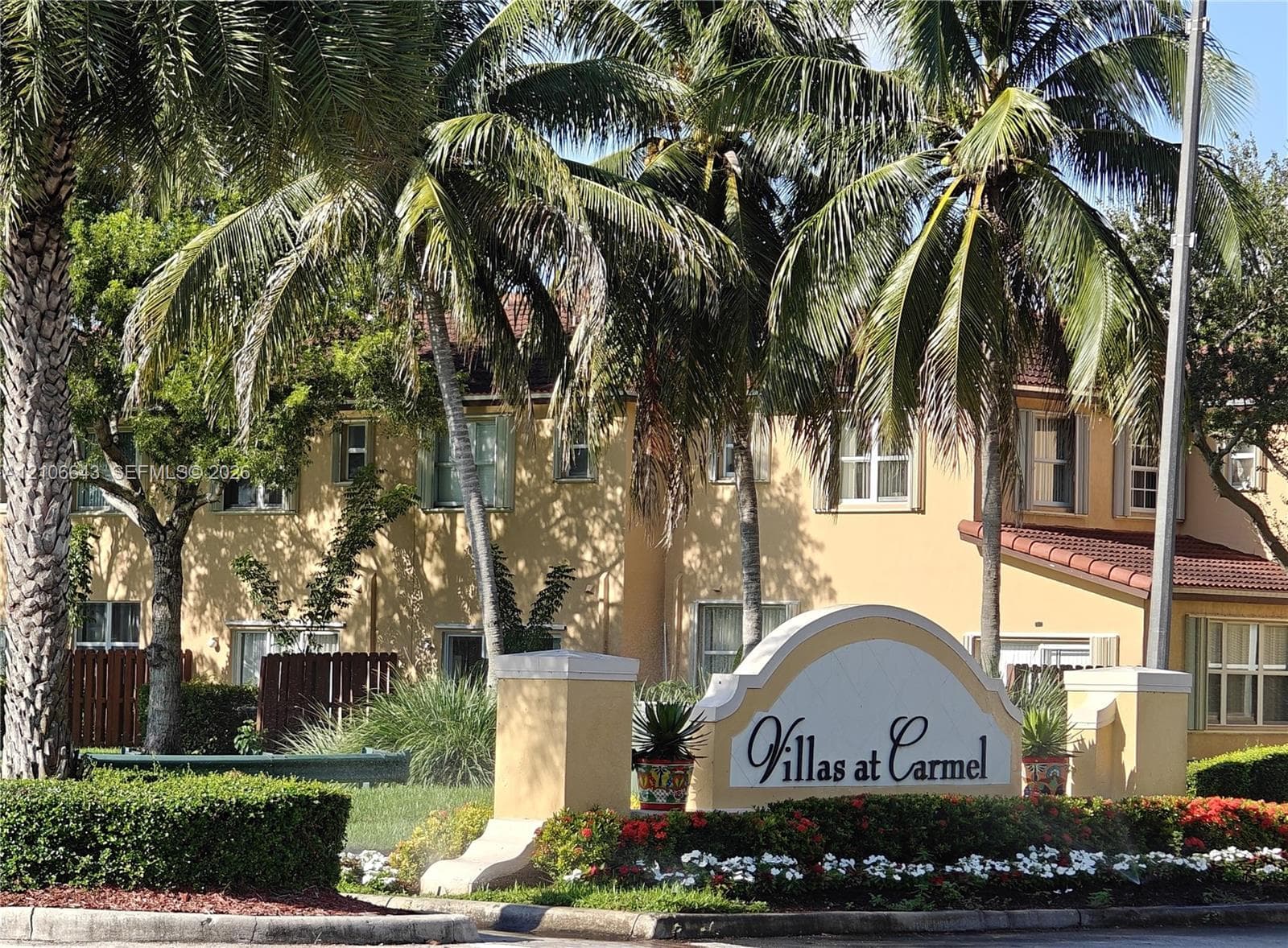 Stucco exterior facade with tiled roof, set within landscaped grounds featuring established palm trees, ornamental shrubbery, and stone-finish signage