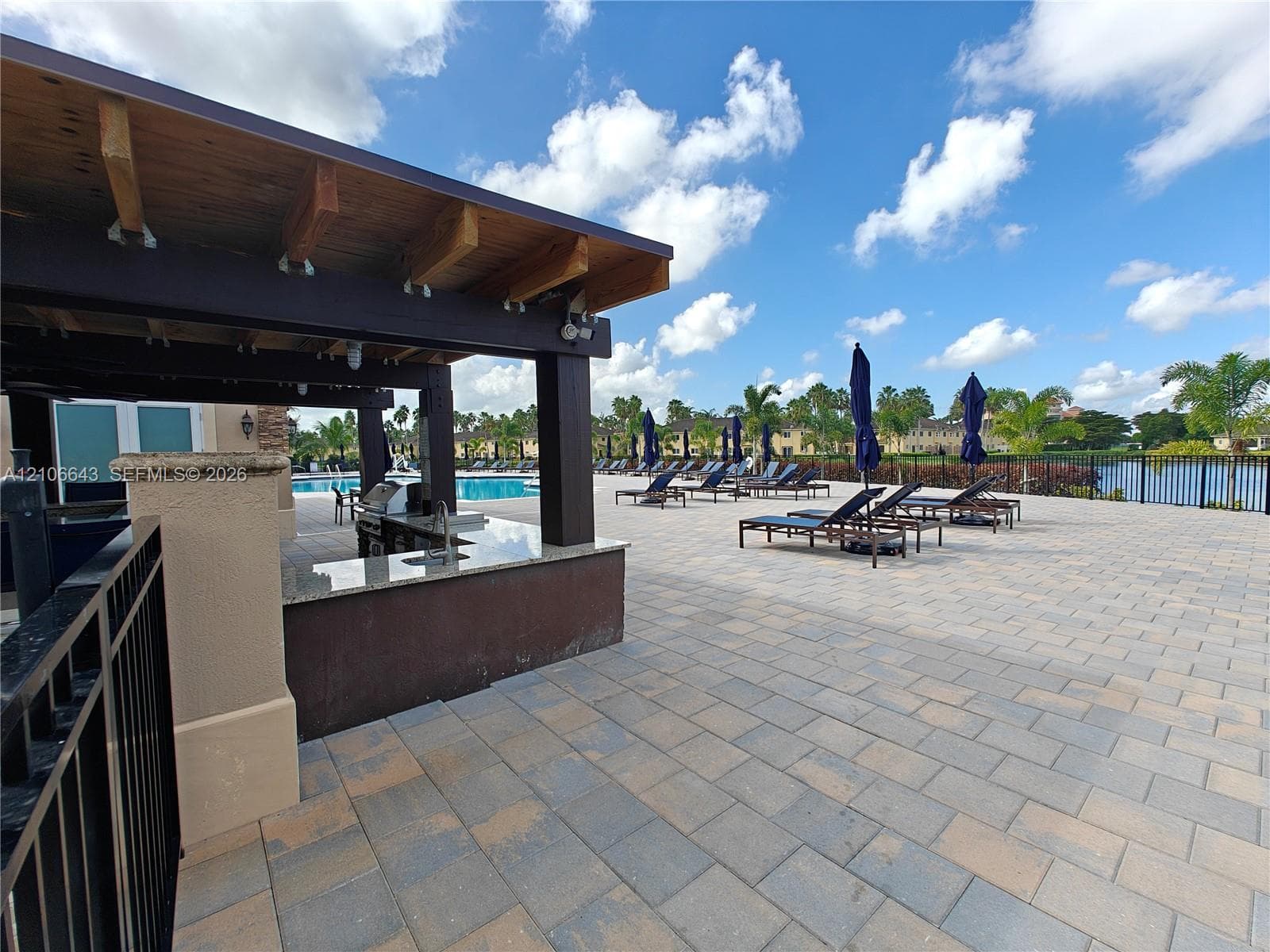 Outdoor kitchen featuring a stone-topped counter, covered timber-frame pergola, expansive paver patio, metal perimeter fencing, and a pool