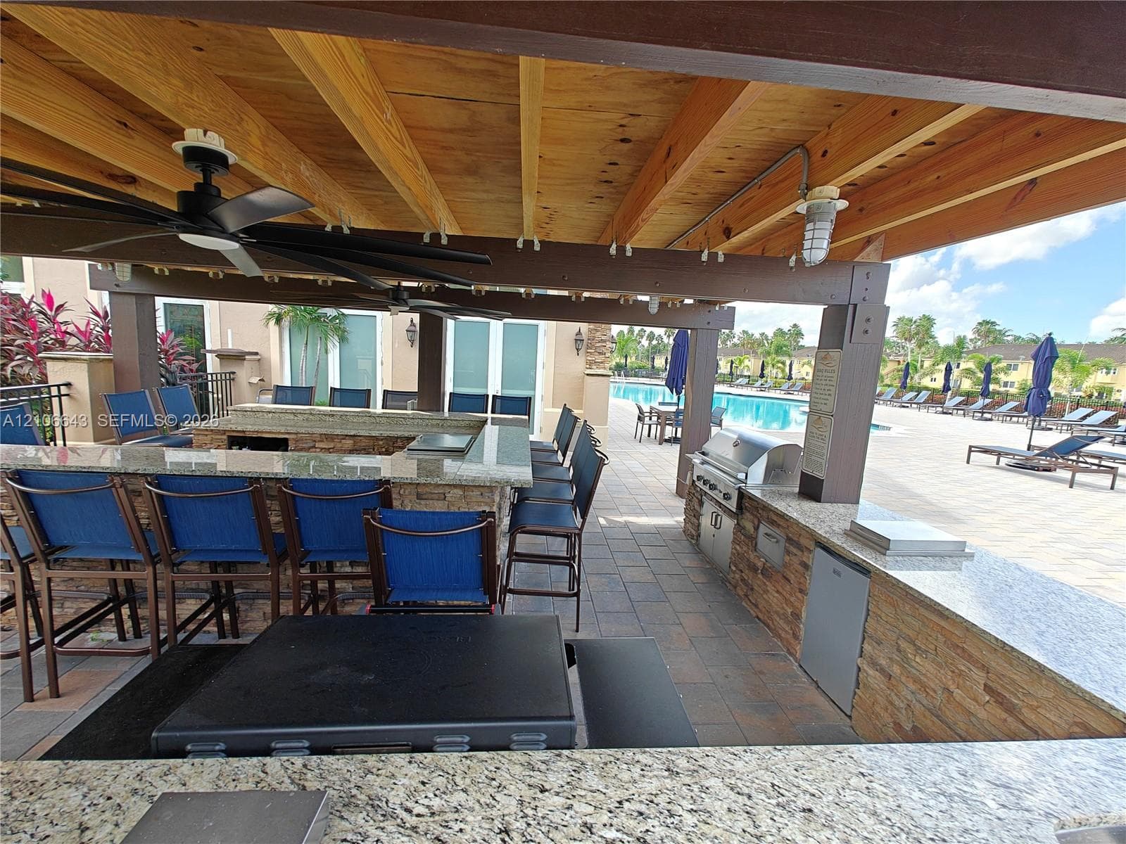 Outdoor kitchen featuring a stone-clad bar, built-in grill, stone countertops, wood-beam ceiling, and ceiling fan