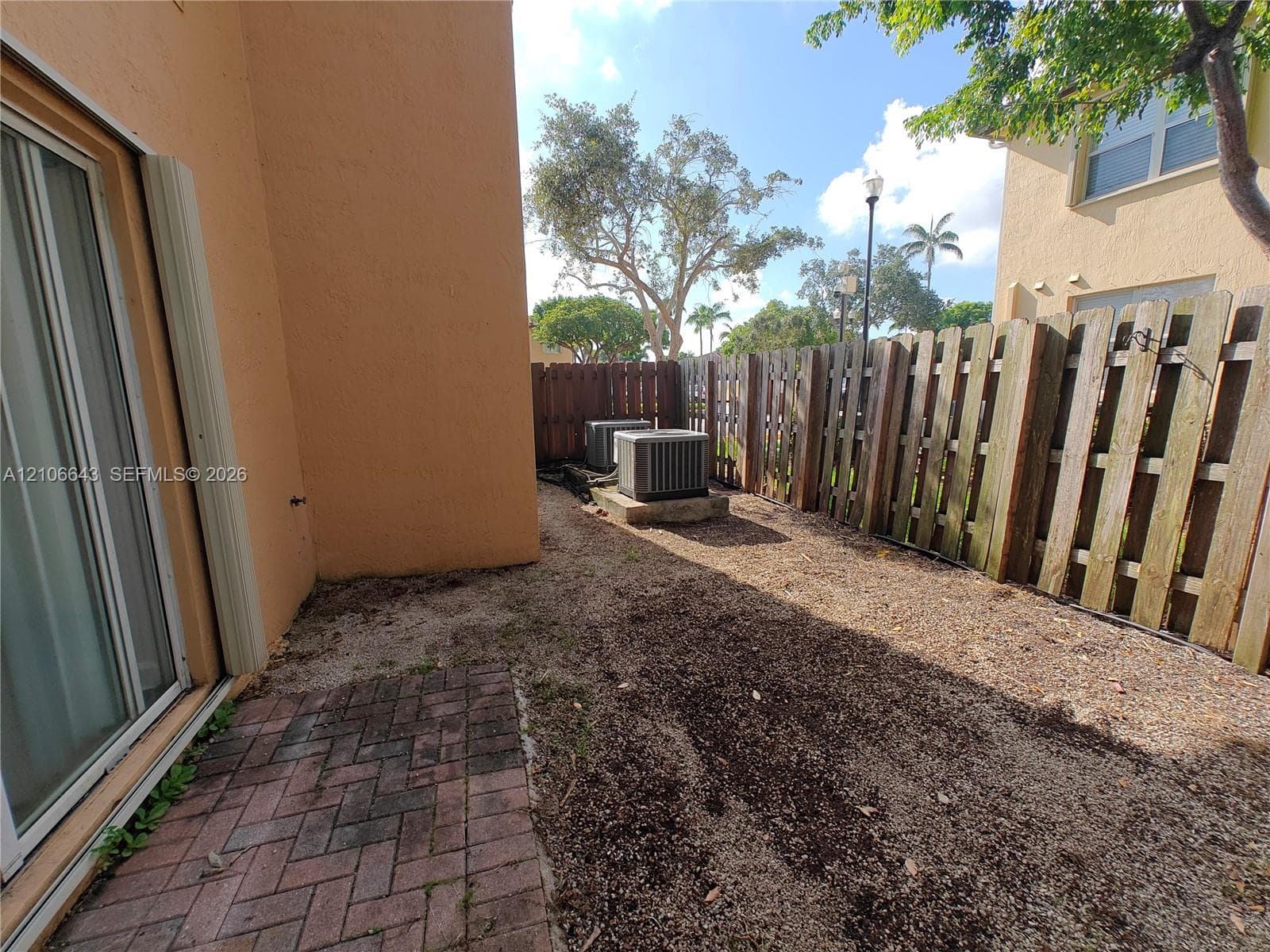 Exterior side yard featuring a herringbone-patterned brick paver patio, wood picket fencing, and two exterior HVAC units on concrete pads