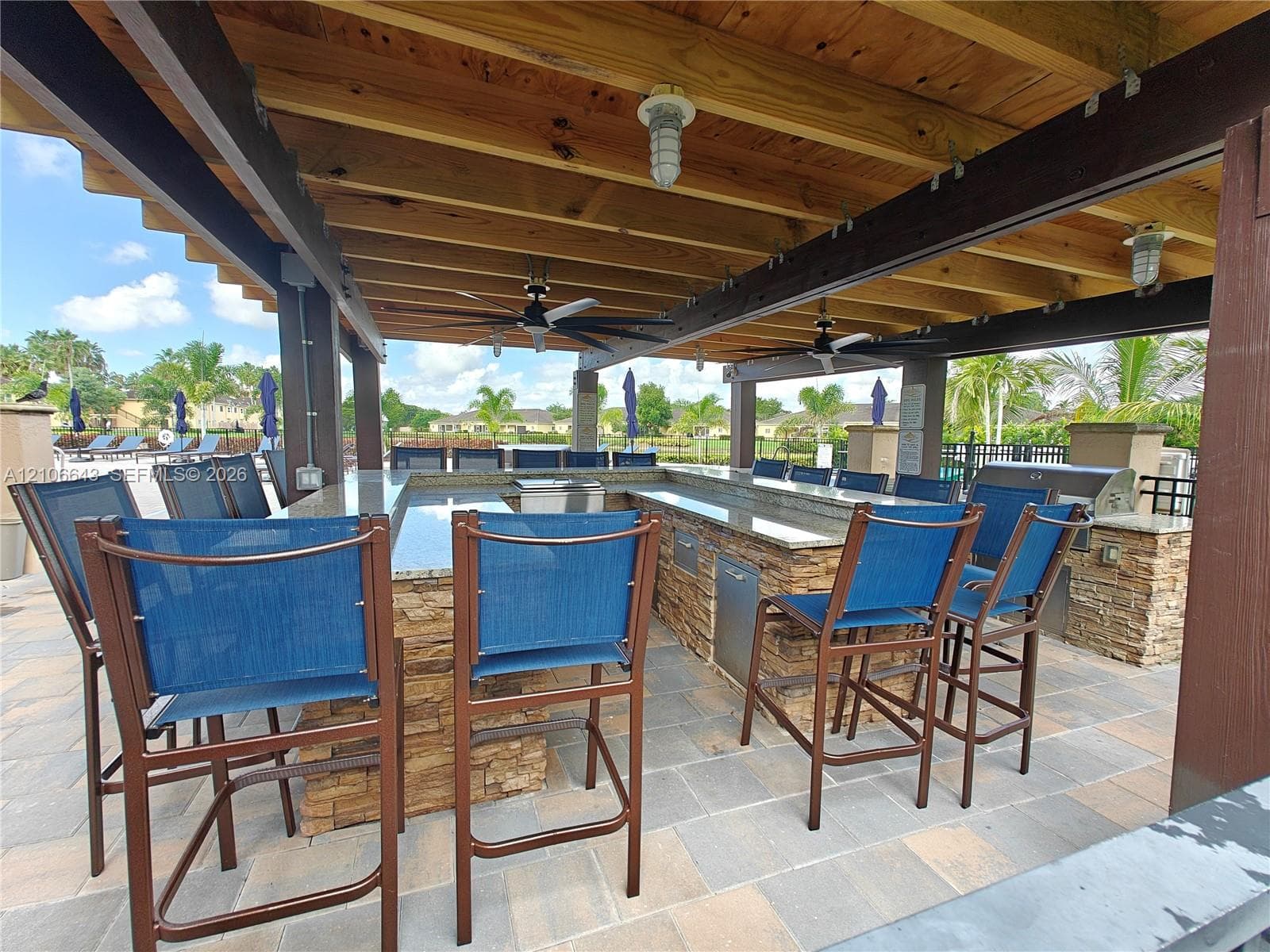 Covered outdoor kitchen featuring a stone-clad bar, stone-veneer base, integrated stainless steel appliances, timber beam ceiling structure, and paver flooring