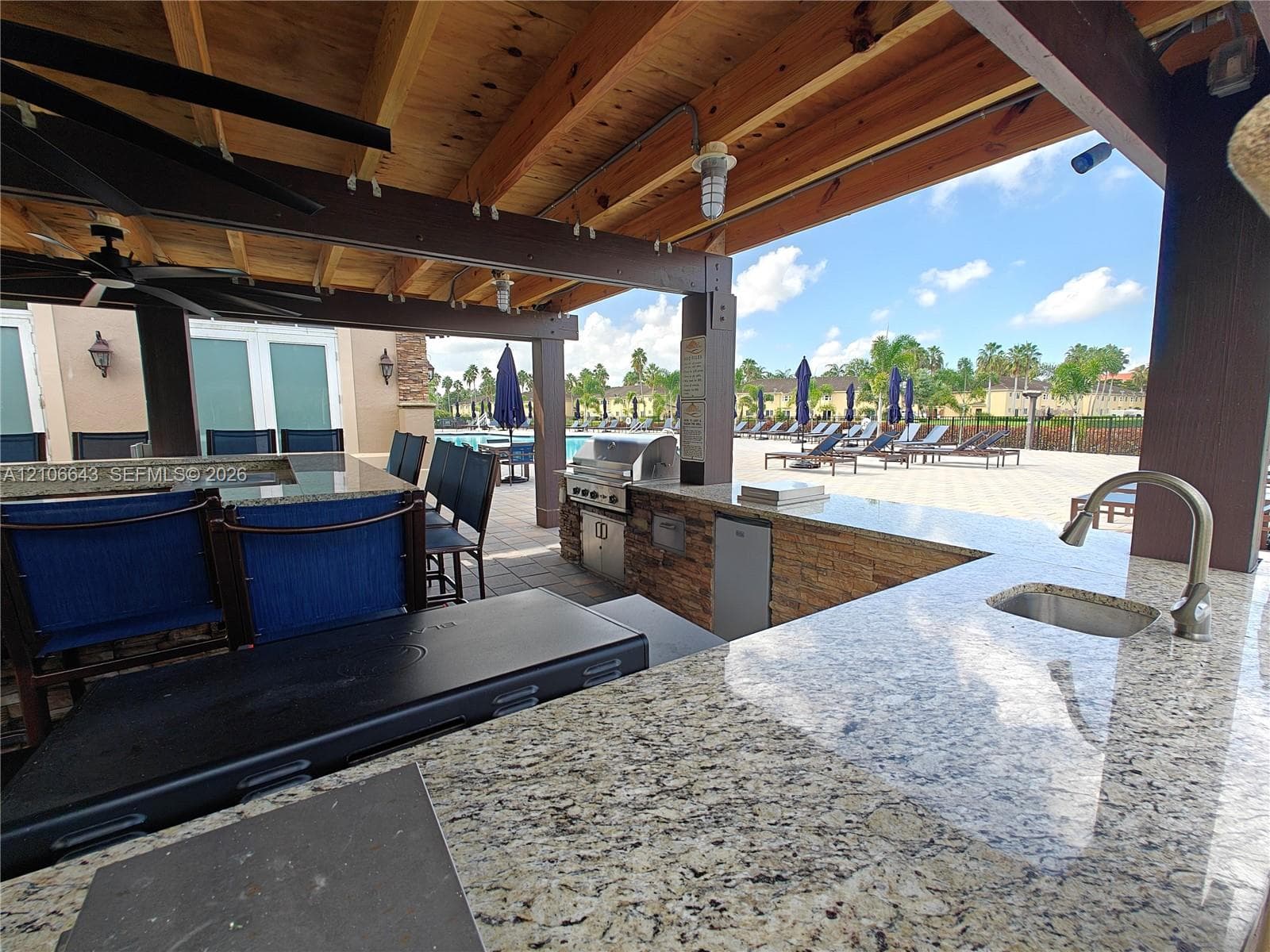 Covered outdoor kitchen featuring a stone-clad base, stone-surface countertops, built-in stainless steel grill, and an undermount sink with a gooseneck faucet