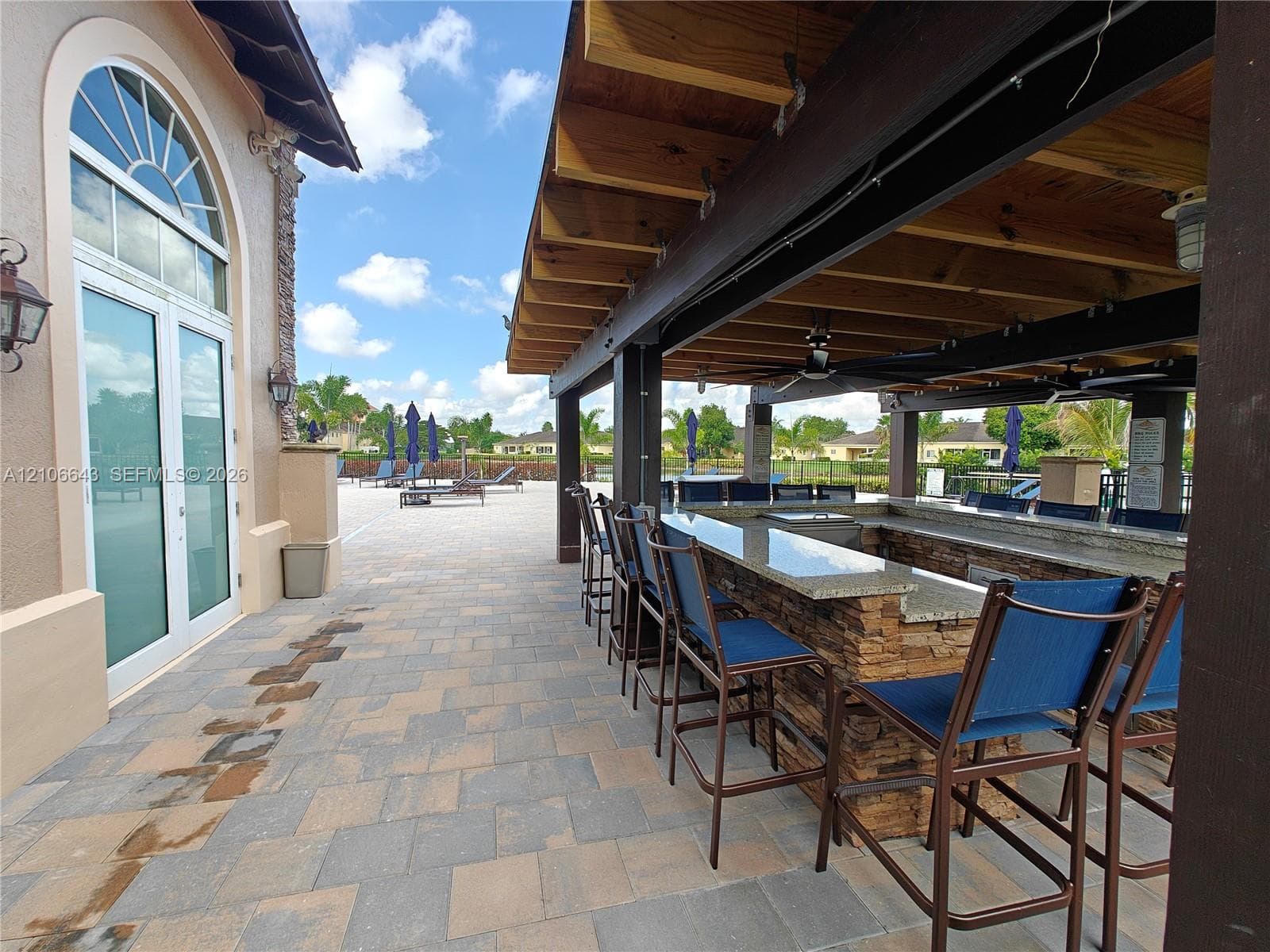 Covered outdoor kitchen featuring a stone-clad bar, solid surface countertops, and an exposed beam wood ceiling, complemented by expansive paver patio hardscaping