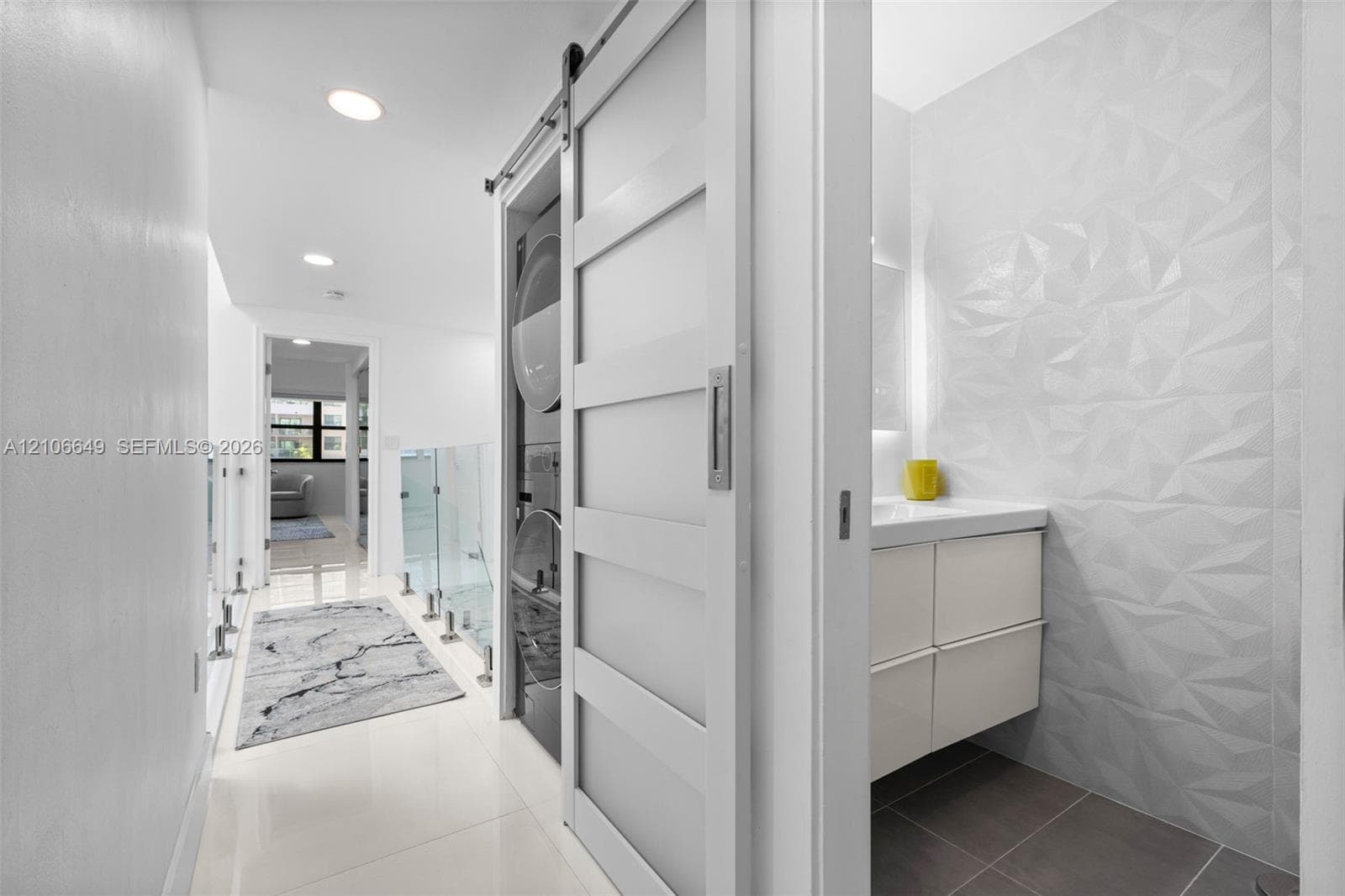 Sliding door entry concealing stacked laundry appliances, textured geometric wall tiling, modern wall-mounted vanity, and glass panel railing