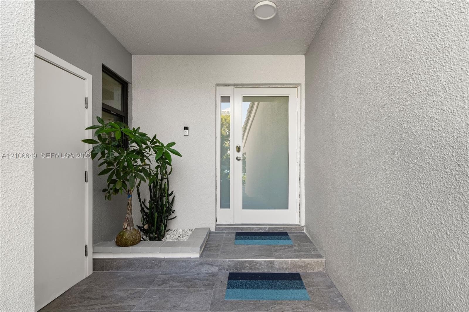 Entryway featuring a white door with a frosted glass panel, recessed ceiling lighting, textured wall finish, grey tiled flooring, and a built-in stone planter