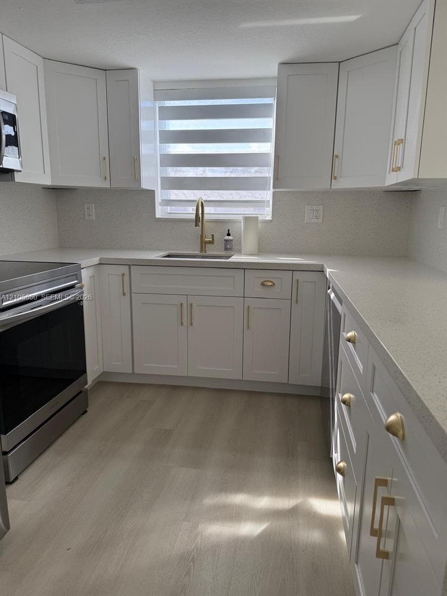 Kitchen featuring white shaker-style cabinetry, quartz countertops, gold-tone hardware, stainless steel appliances, and wood-finish flooring