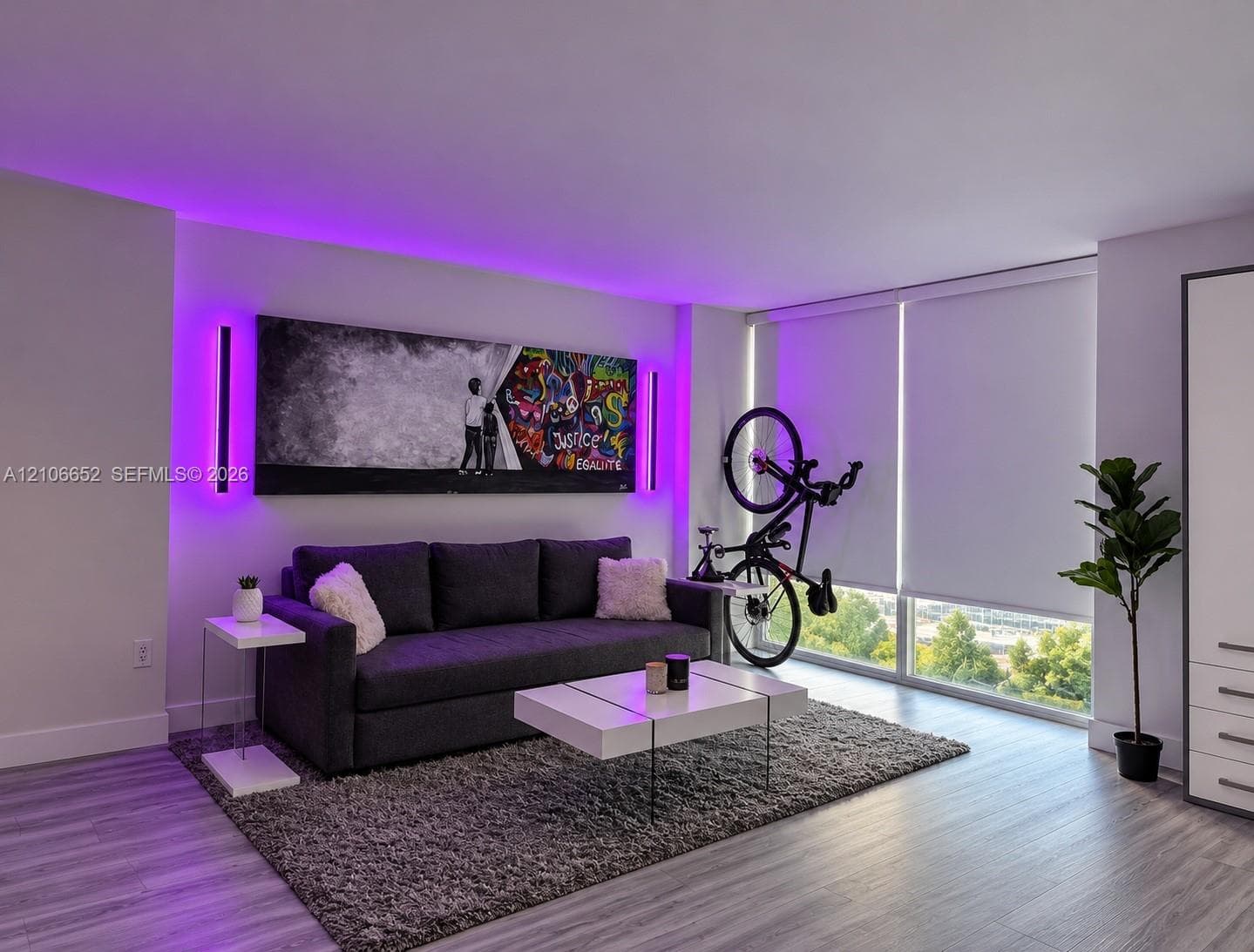 Living area featuring wood-finish flooring, floor-to-ceiling windows with roller shades, integrated cove lighting, and dual vertical wall-mounted fixtures