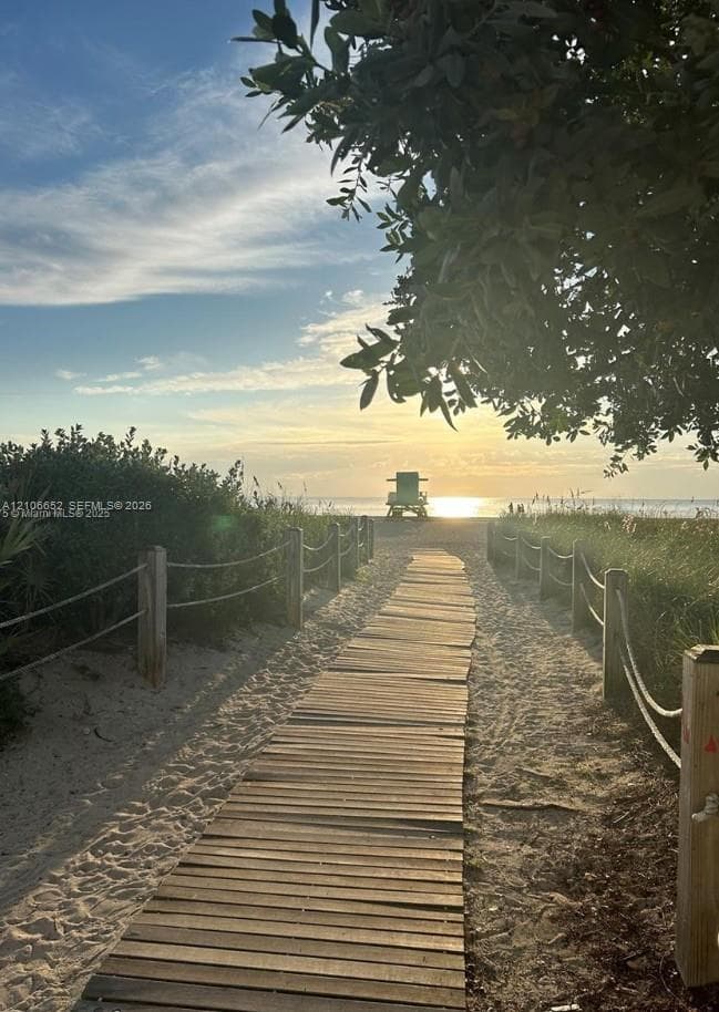 Wood-slat boardwalk with rope-and-post fencing leading to sandy coastal frontage, framed by mature overhanging foliage in rear building leading to BEACH