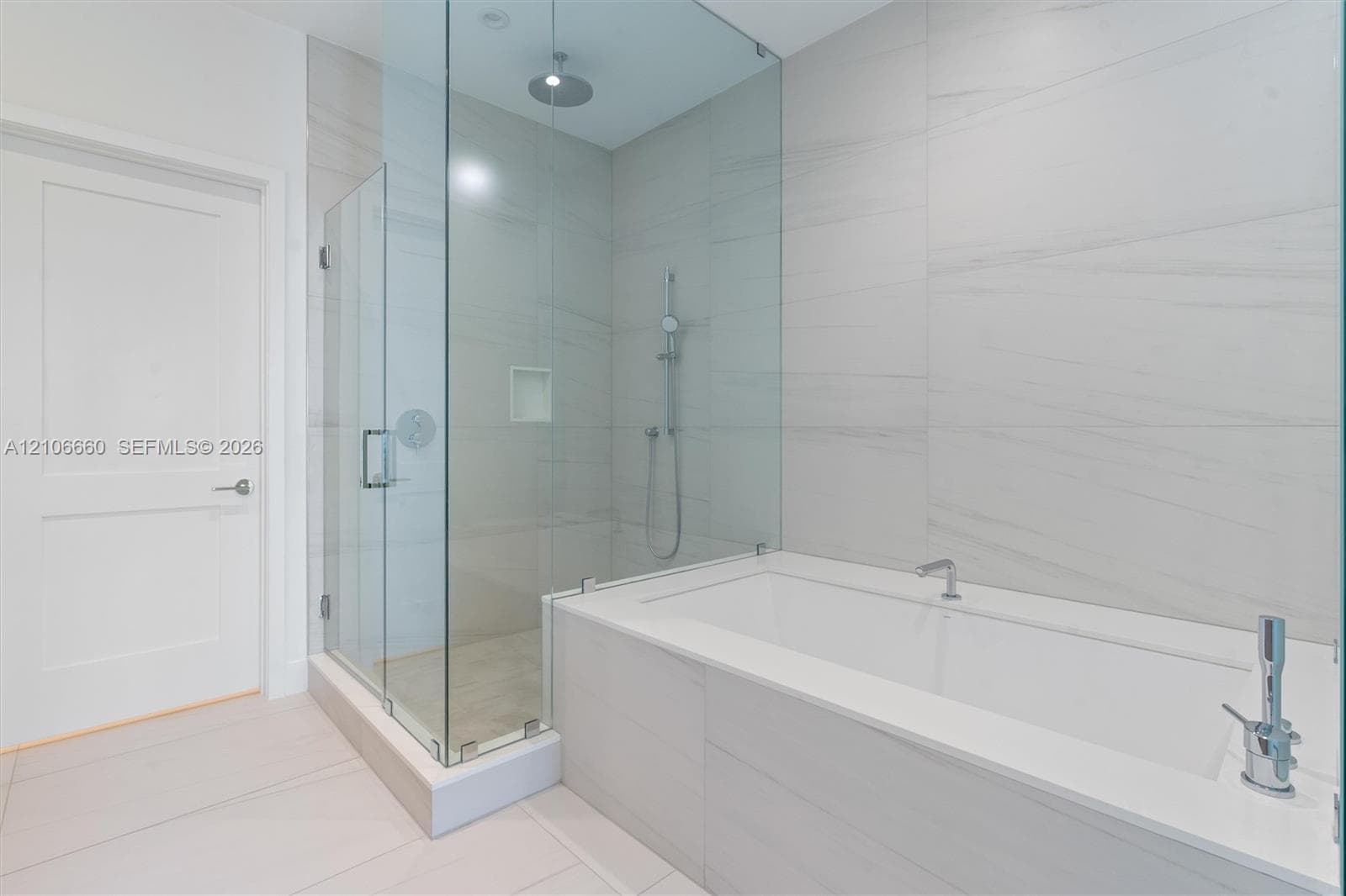 Walk-in glass-enclosed shower featuring a rainfall showerhead, integrated tiled wall niche, deep soaking tub with deck-mounted faucet, and large-format porcelain wall tiling