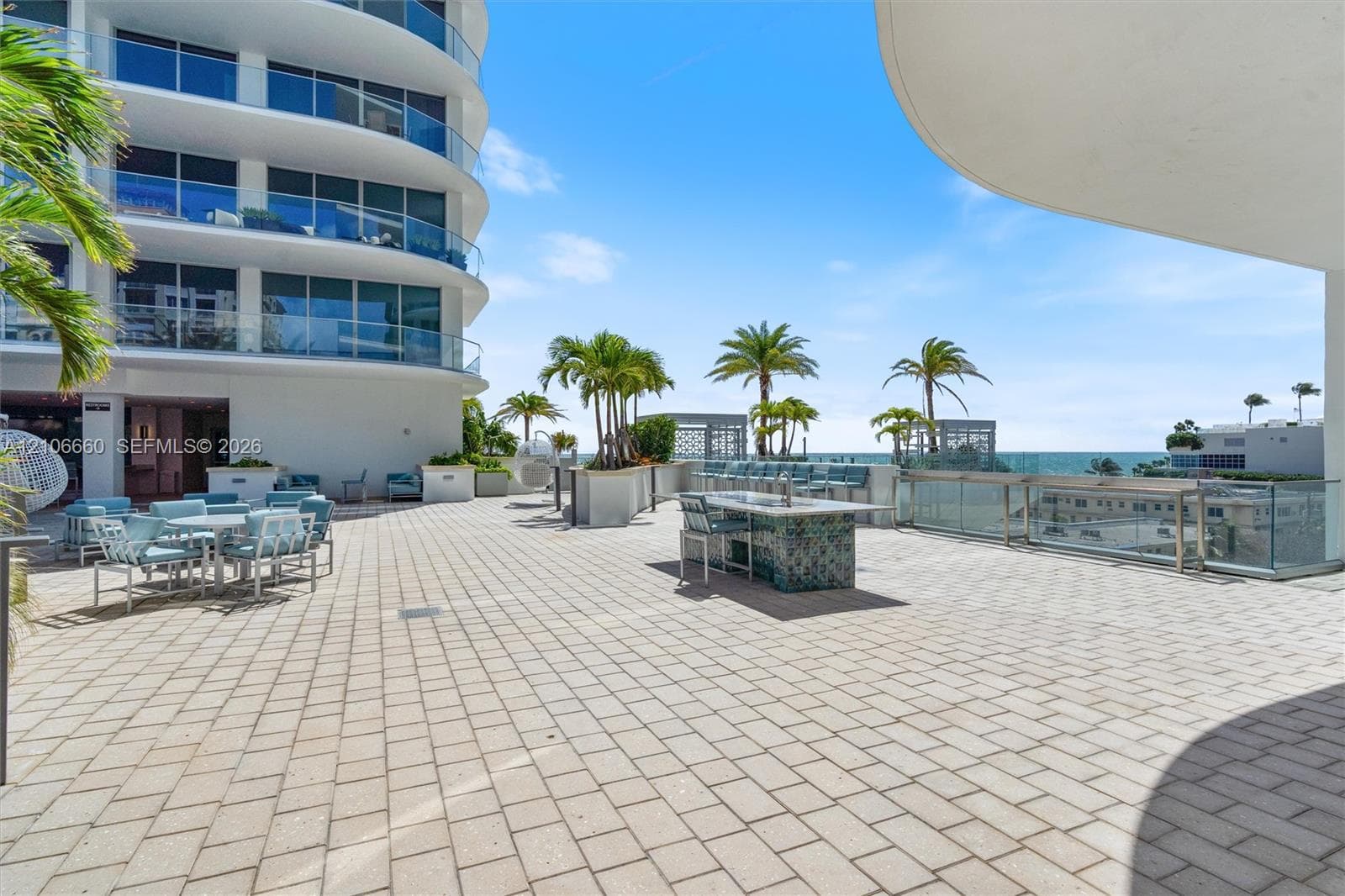 Expansive paved terrace featuring a tiled outdoor kitchen island, glass-panel perimeter railing, and mature palm landscaping