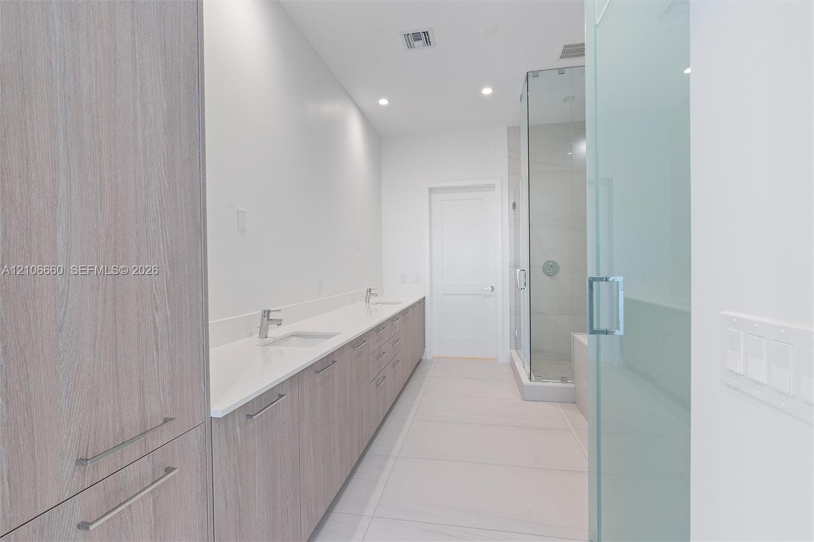 Dual-sink vanity with wood-finish cabinetry, light stone countertop, walk-in glass-enclosed shower, and tile flooring