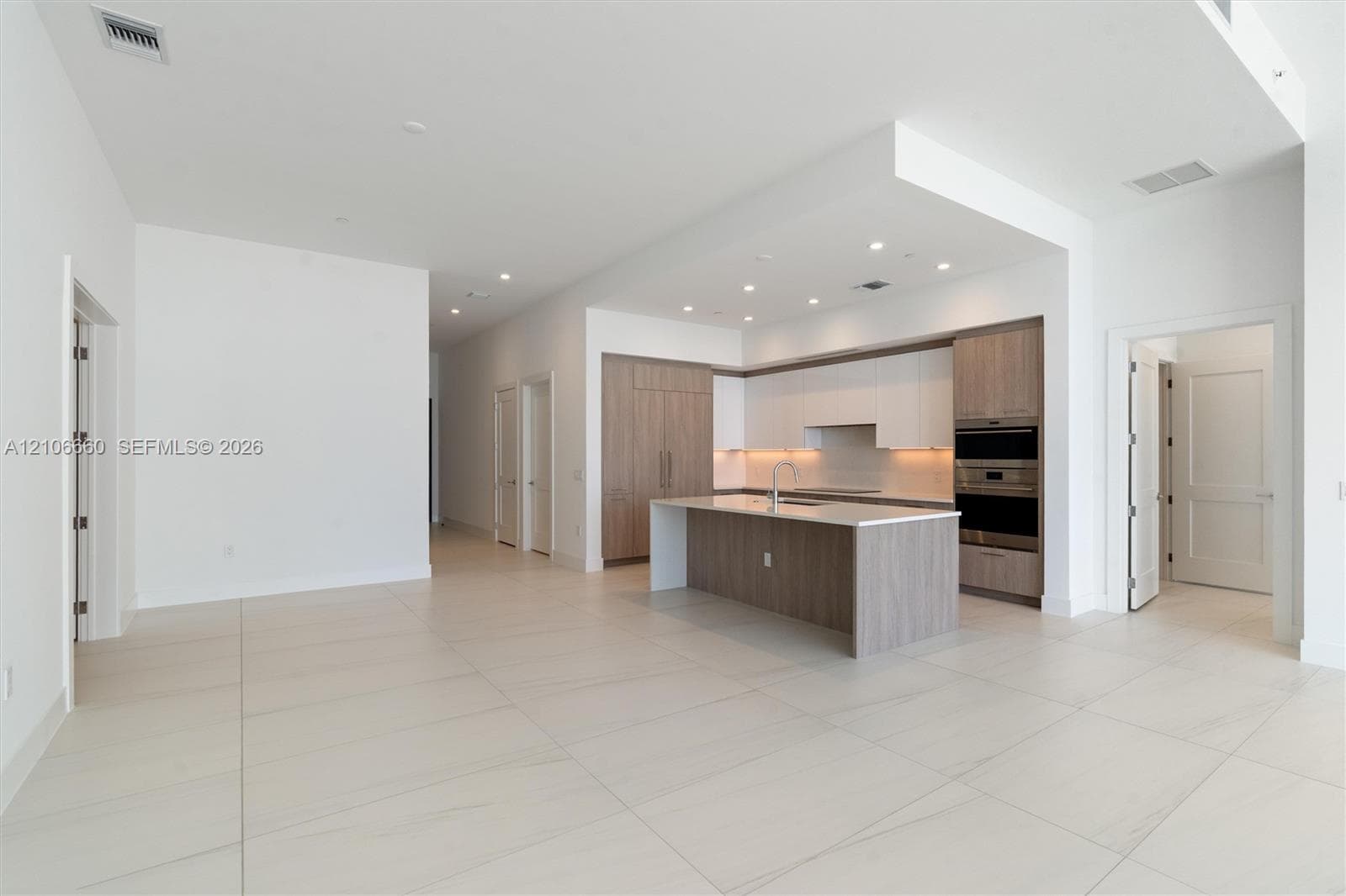 Open-concept kitchen featuring two-toned cabinetry, stone-finish countertops, a central island with an integrated sink, built-in stainless steel appliances, and large-format tile flooring