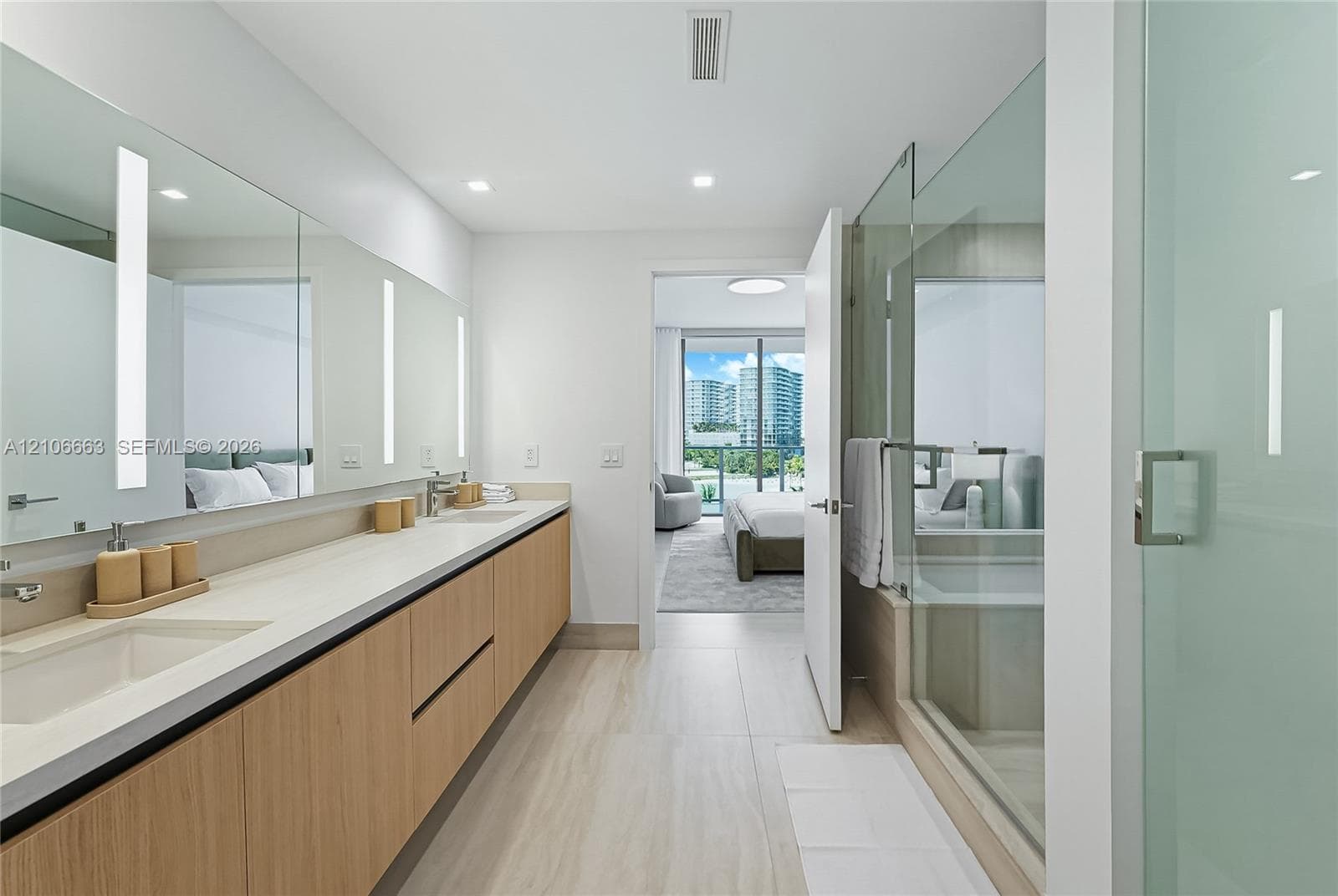 Dual-sink vanity with light wood-finish cabinetry, solid-surface countertop, full-width vanity mirror with integrated vertical lighting, and glass-enclosed shower with tile flooring